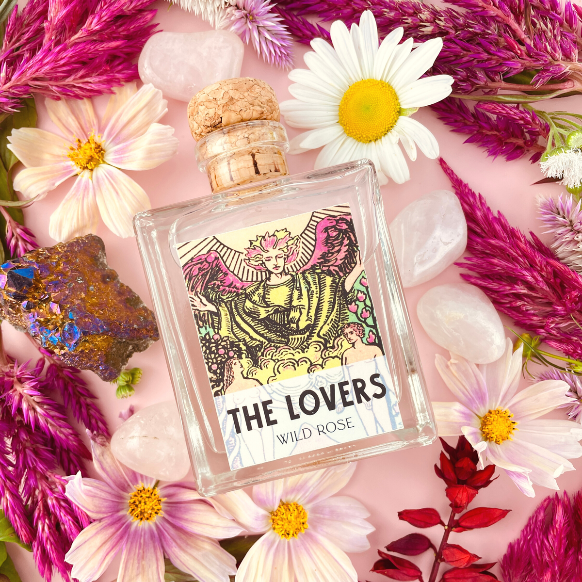 Perfume bottle labeled 'The Lovers' by Wild Rose surrounded by flowers and crystals on a pink background