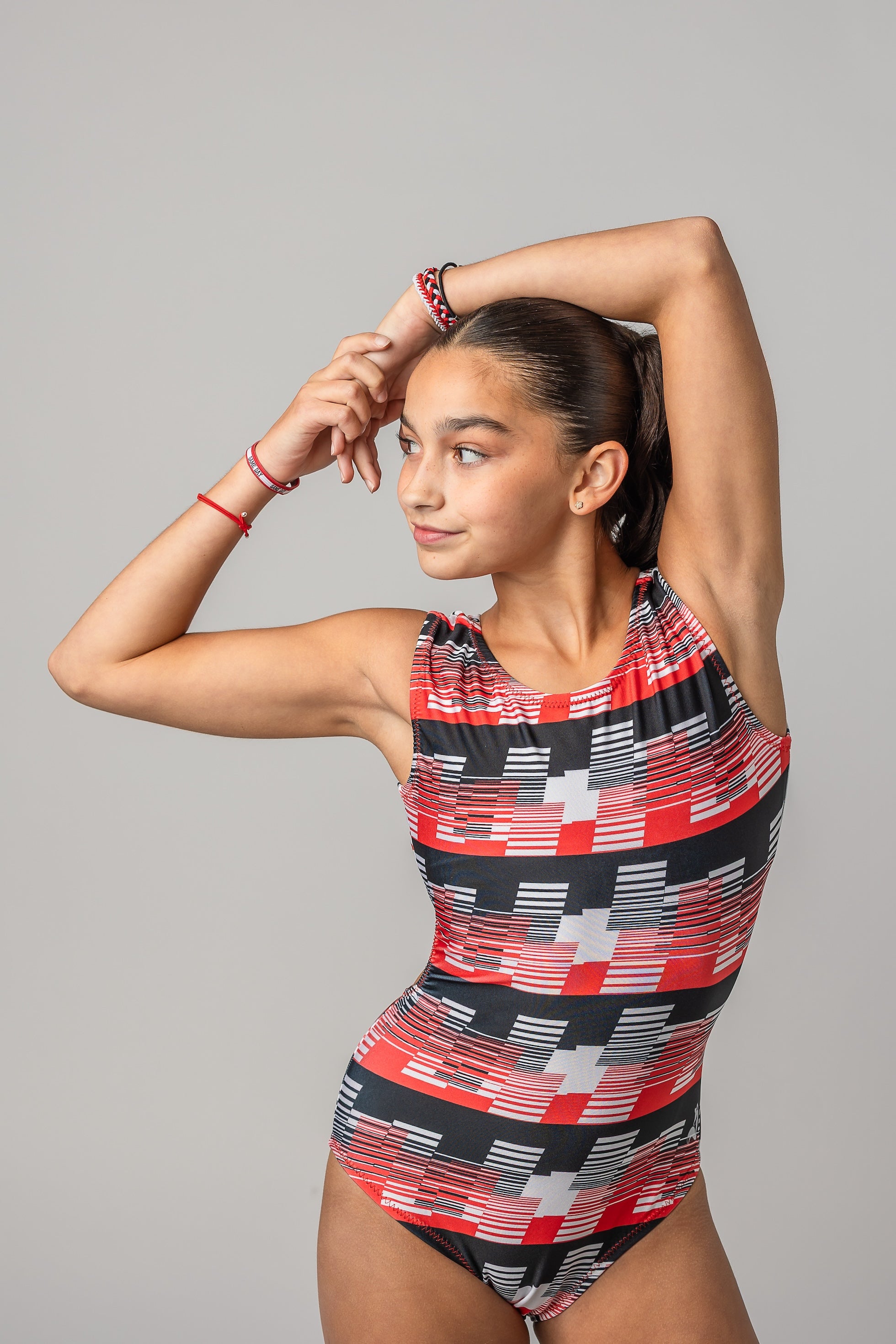 A young female gymnast modeling a sleeveless leotard for girls with a red, black, and white geometric pattern, posing against a solid gray studio background.