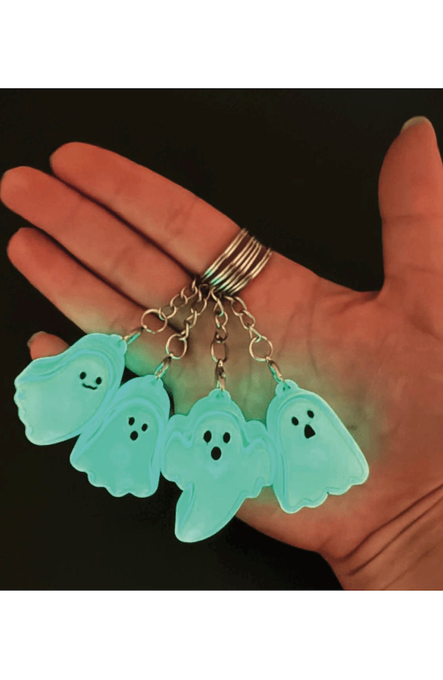 Four glowing mint green ghost clicker keychains with black facial features, attached to a silver metal ring, held in a person's hand against a dark background.