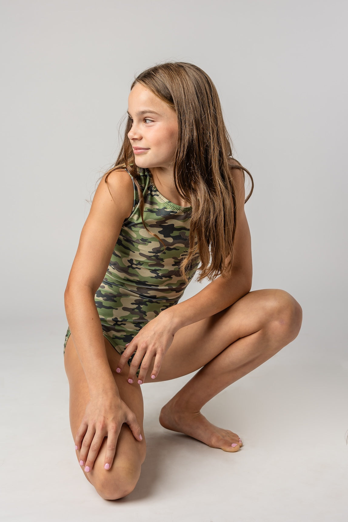 A young girl with long brown hair crouches in a studio setting, wearing a sleeveless camouflage-print leotard against a light gray background.