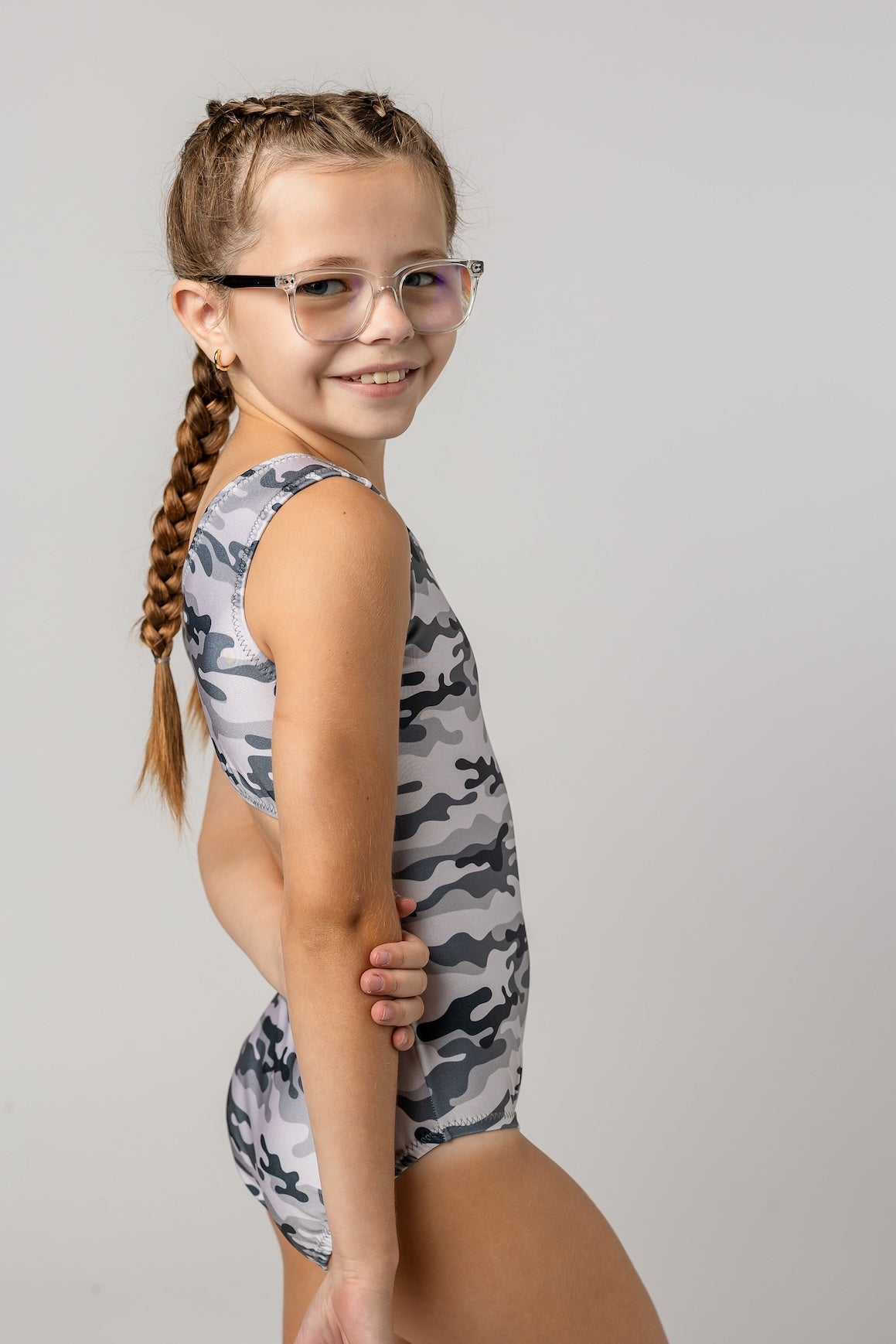 A young girl with glasses and a long braided ponytail is wearing a sleeveless leotard for girls with a gray and white camouflage pattern, posing against a light gray studio background.