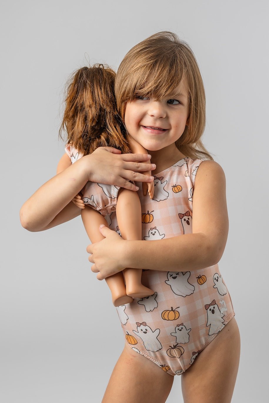 A young girl with light brown hair smiles while hugging a doll. She is wearing a sleeveless one-piece toddler gymnastics leotard set in a light peach and white gingham pattern with a print of white ghosts and orange pumpkins, against a solid light gray studio background.