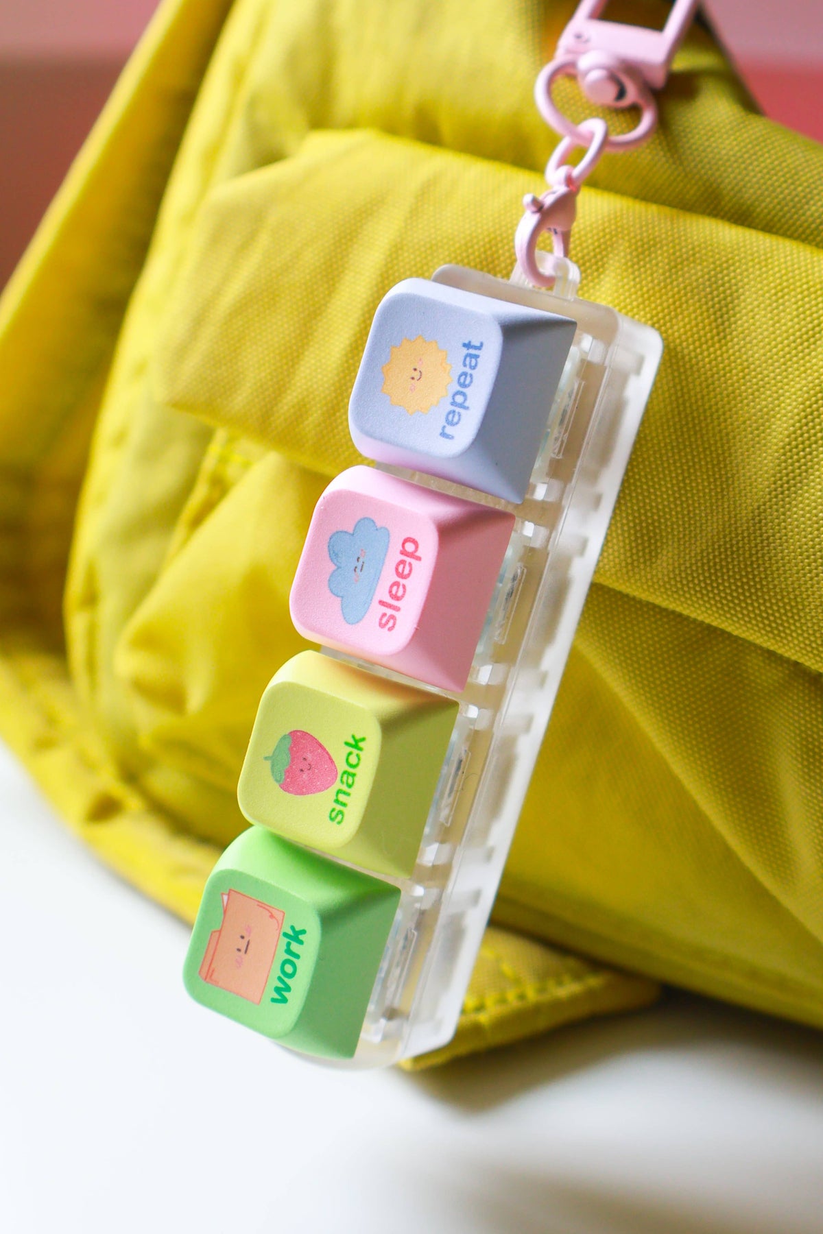 A pastel-colored keycap charm with four labeled keys reading repeat, sleep, snack, and work, attached to a clear plastic clip and hanging near a bright yellow fabric bag.