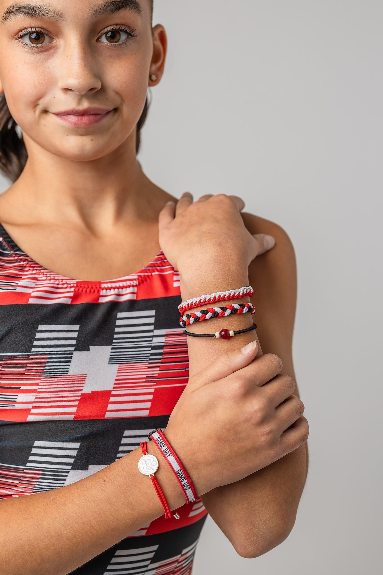 A close-up of a girl wearing several colorful braided and cord bracelets on her wrist, including red, white, and black designs, against a light gray background.