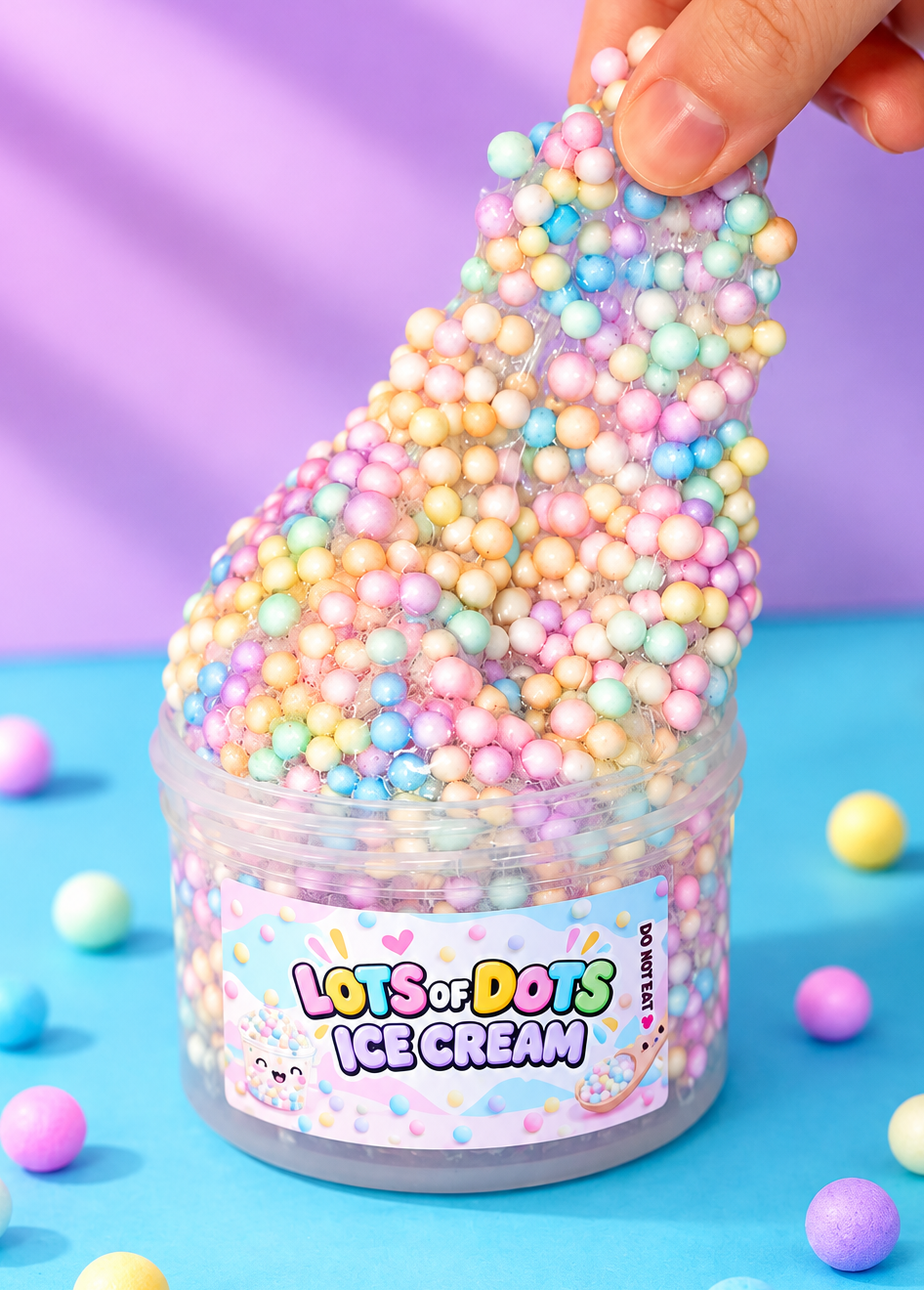 A hand stretches a pile of pastel foam beads slime inside a clear plastic jar labeled 'Lots of Dots Ice Cream' against a purple and blue background.