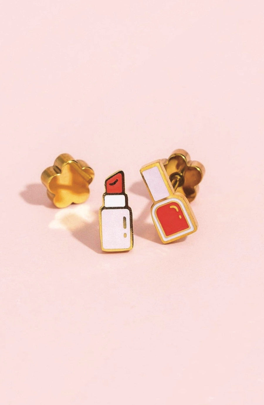 Three gold-tone enamel pins shaped like a flower, lipstick, and an open compact with red and white details, arranged on a soft pink background.