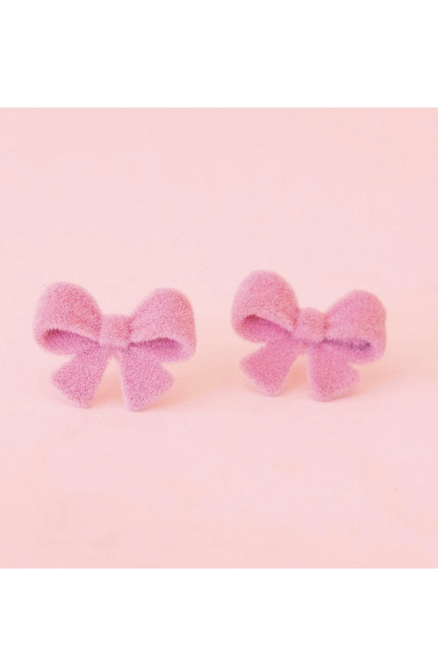 A pair of small fuzzy bow-shaped stud earrings in a soft pastel pink-purple color, displayed on a solid light pink background.