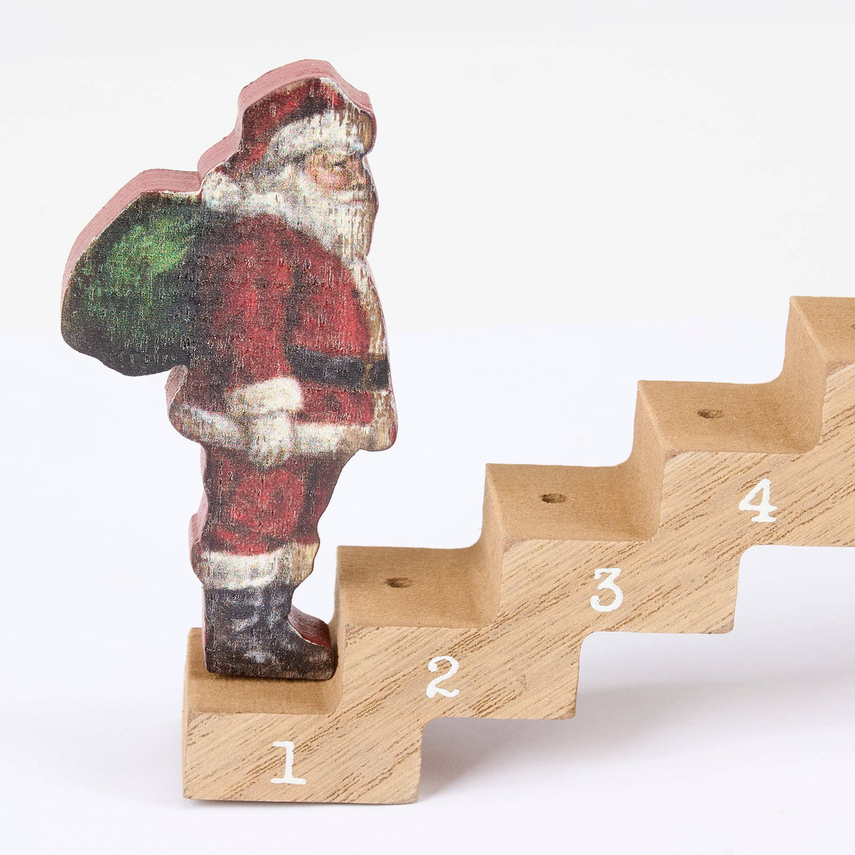 A small wooden Santa Claus figurine wearing a red suit and carrying a green sack, standing on a numbered wooden step pyramid against a plain white background.