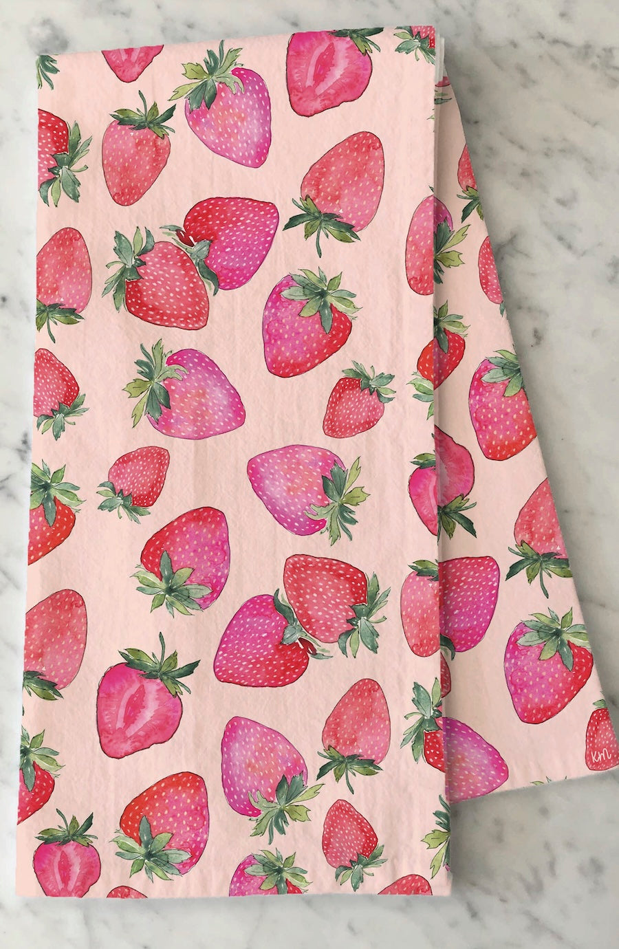 A folded light pink fabric item, likely a tea towel or napkin, featuring a watercolor-style print of red strawberries with green leaves, displayed on a white marble surface.