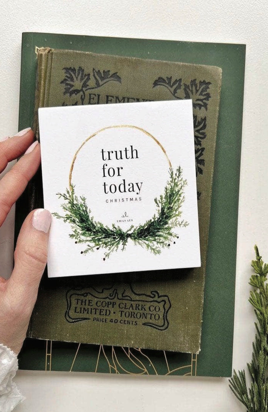 A hand holds a white Christmas greeting card that reads 'truth for today CHRISTMAS' with a green wreath illustration, layered over an old green book.