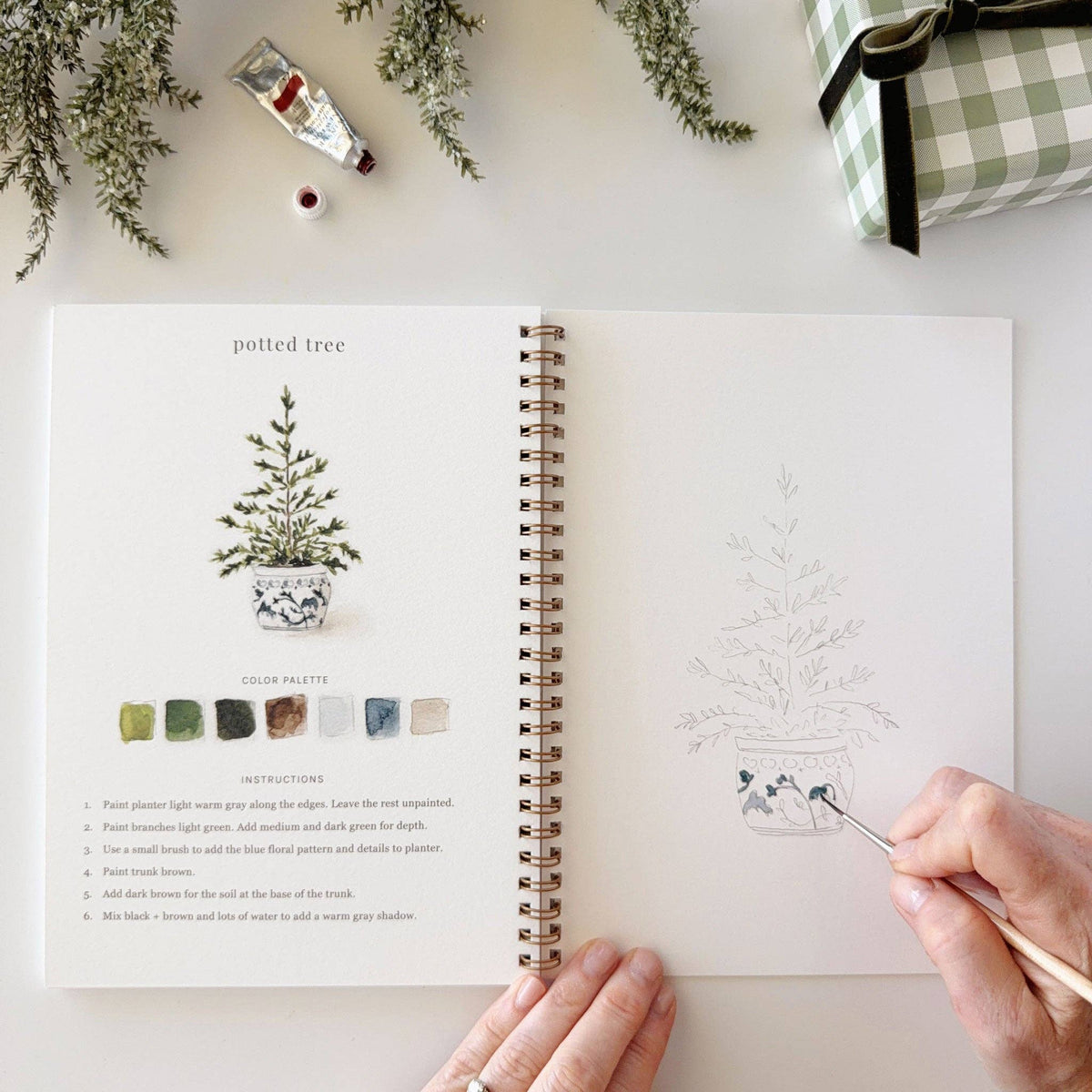 A spiral-bound art journal or coloring book shown open on a white surface, with a hand holding a paintbrush near the illustrated pot tree page. The left page has a watercolor potted tree, color swatches, and instructions, while the right page has a lightly sketched version of the same design.