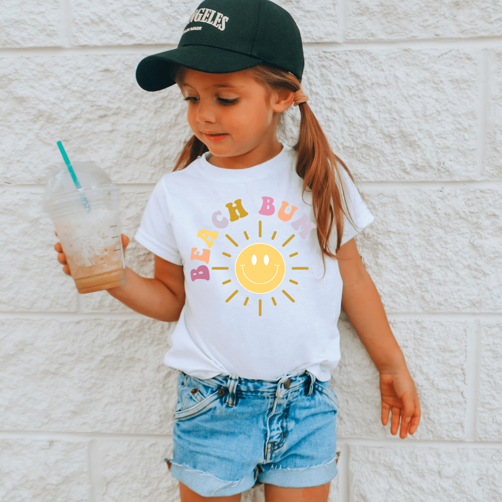 Girl in 'Beach Bum' white t-shirt with sun graphic and denim shorts.