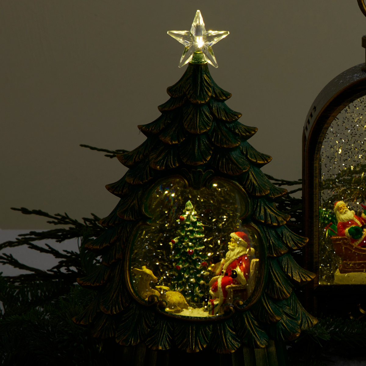 A decorative Christmas tree figurine with a glowing star on top and a snow globe scene inside, featuring Santa Claus, a small Christmas tree, and a reindeer. Part of another similar ornament is visible on the right against a dark background.