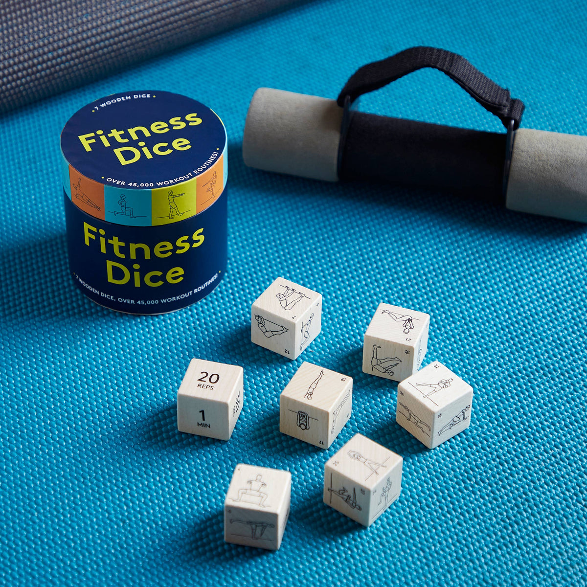 Fitness dice set with a navy blue cylindrical tin labeled 'Fitness Dice,' several off-white polyhedral dice with printed exercise icons, a foam roller, and a black carrying strap on a bright blue textured mat.