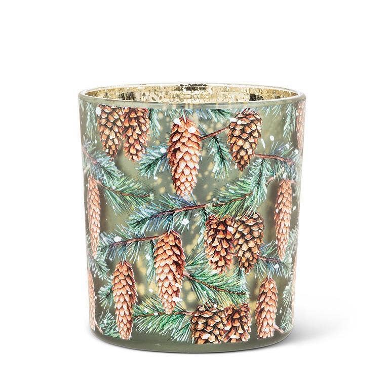 A cylindrical glass candle jar featuring a printed winter pine design with brown pinecones and green pine needles on a muted olive background, with a metallic gold interior rim.