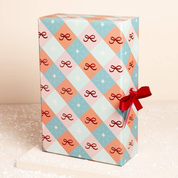 A rectangular gift box wrapped in holiday-themed wrapping paper with a light blue, peach, and pink argyle pattern featuring dark red bows and white snowflake motifs, tied with a red ribbon bow on the right side.