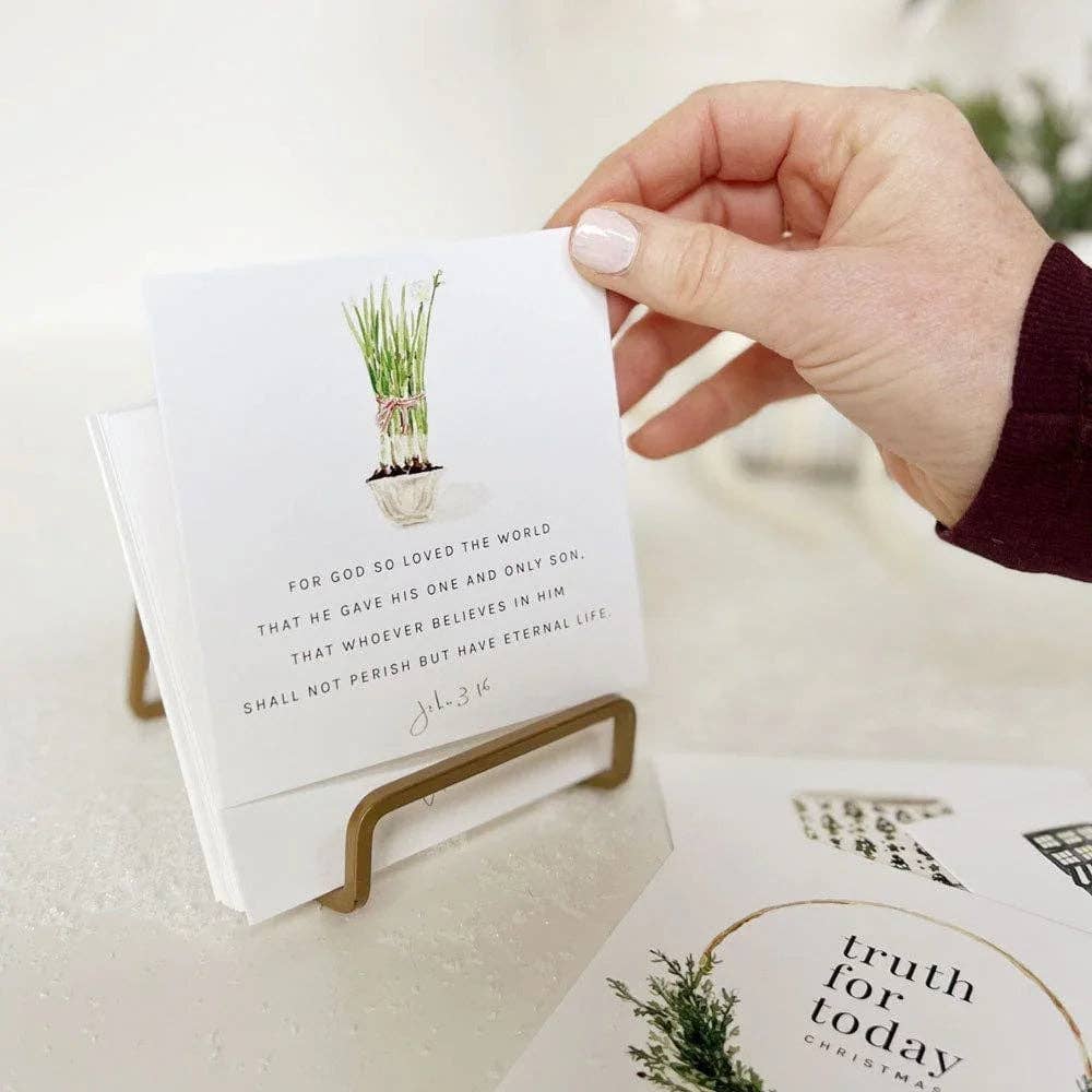 A hand with light pink manicured nails holds a small white greeting card on a gold metal stand. The card features a watercolor illustration of green grass in a pot and a Bible verse from Jeremiah 3:19.