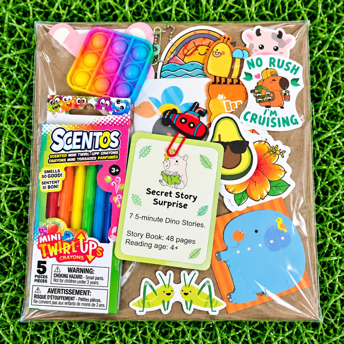 A packaged children's activity set on a green grass-like surface. The clear plastic bag contains a rainbow pop-it toy, a box of Mini Twirl-Up crayons, a 'Secret Story Surprise' card, and assorted dinosaur-themed stickers.