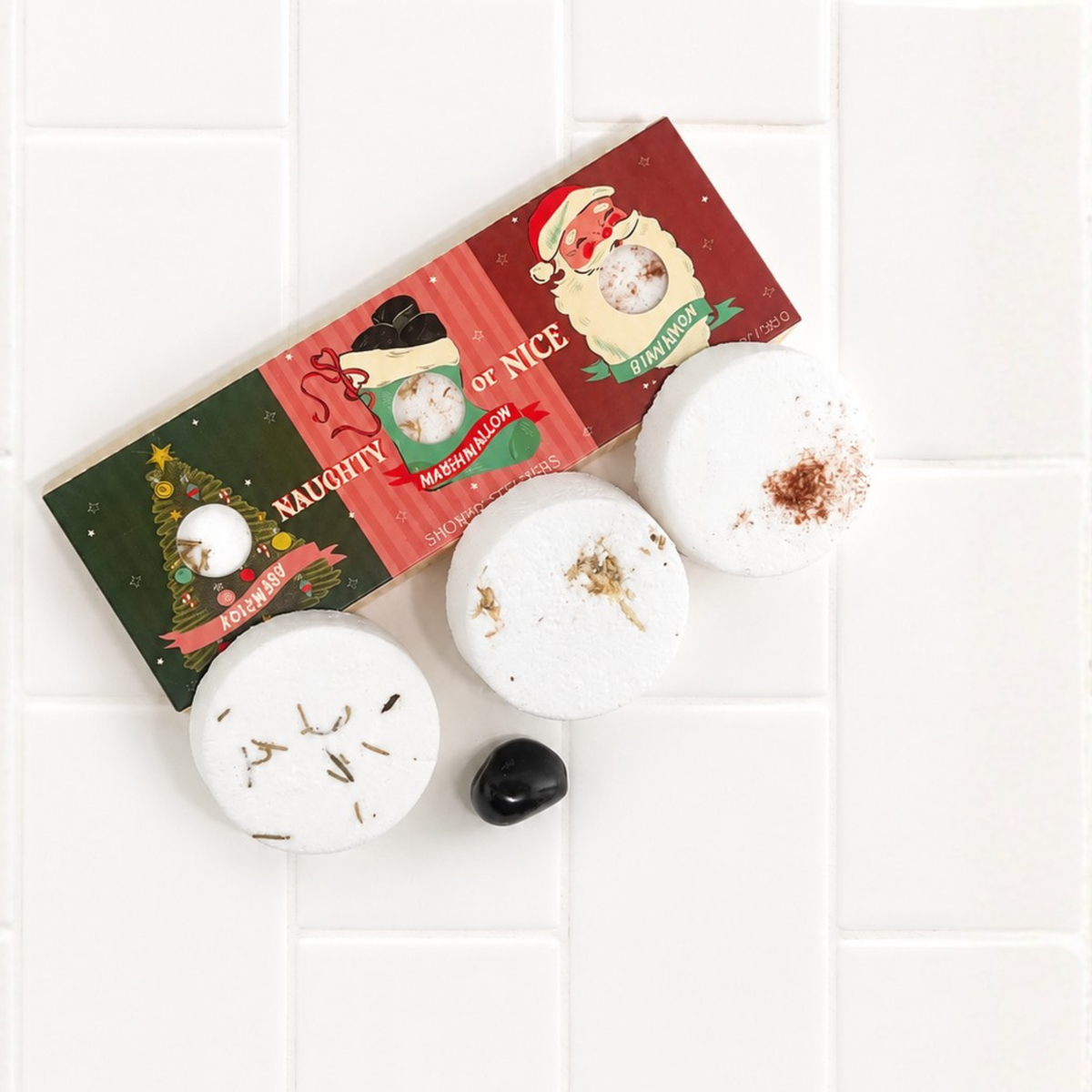 Three round white bath bombs with holiday-themed botanical inclusions are arranged on a white tiled surface next to their illustrated packaging box.