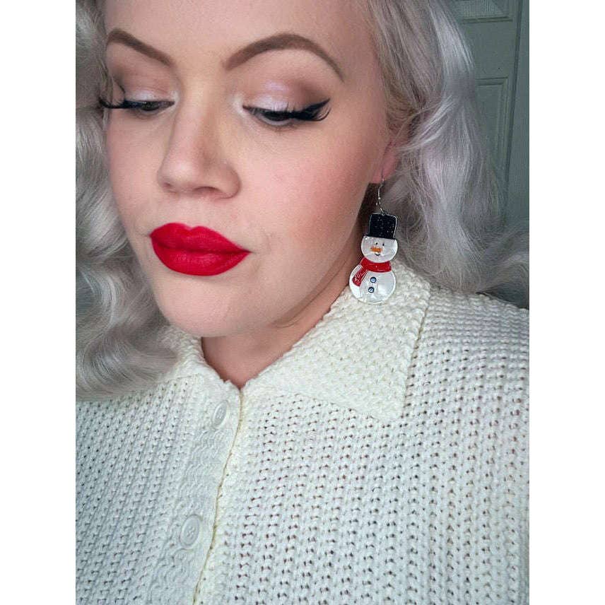 A close-up of a woman with silver hair wearing a cream knit top and a red snowman earring.