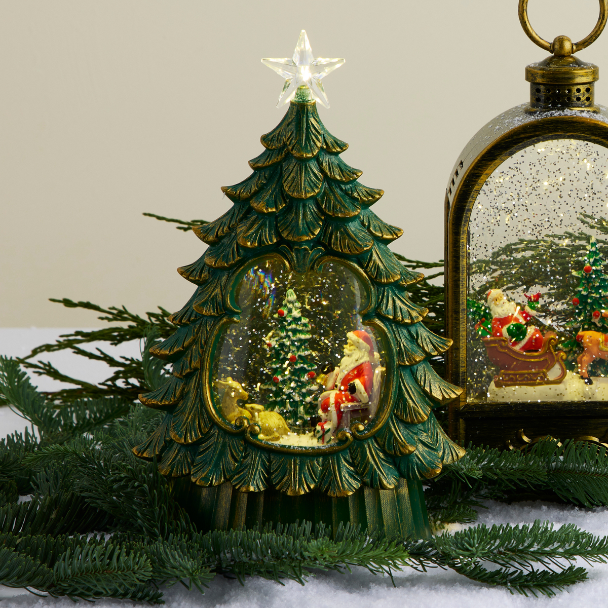 A green Christmas tree snow globe figurine with gold accents and a star topper, shown beside a matching lantern-style ornament on a tabletop with evergreen branches and faux snow.