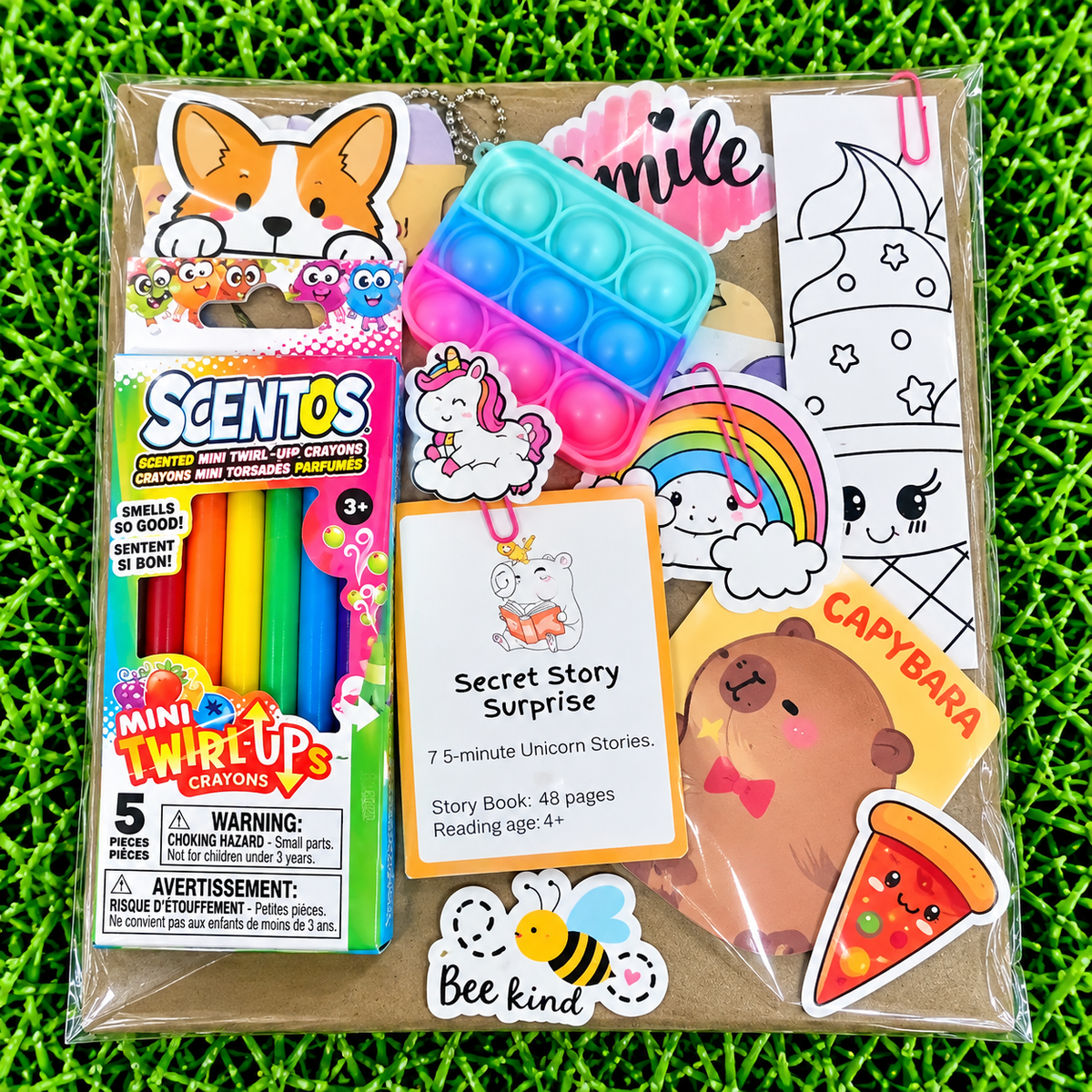 A packaged mini surprise toy set on a bright green grass-like background. The clear plastic bag contains a Scents Mini Twirl-Up Crayons box, a blue and pink pop-it toy, a 'Secret Story Surprise' story card, and several colorful stickers including a corgi, unicorn, rainbow, bee, and pizza slice.