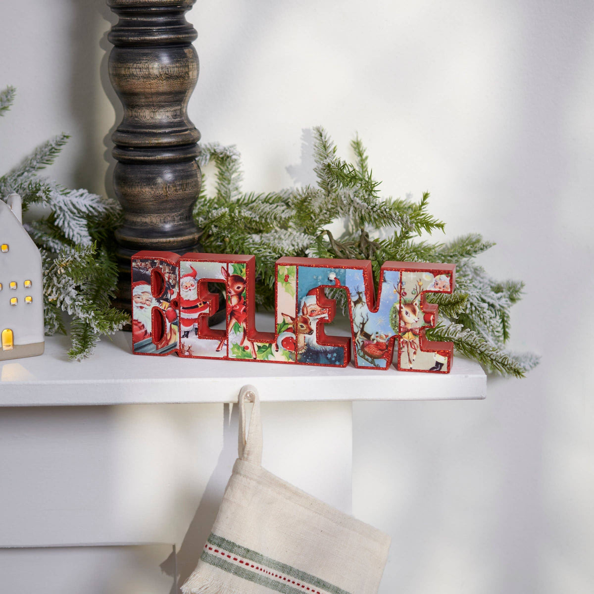 A decorative tabletop scene with a white shelf holding a large red-and-white letter sign spelling "BELIE" surrounded by green garland and a small white house ornament. A cream stocking hangs below the shelf against a white wall.