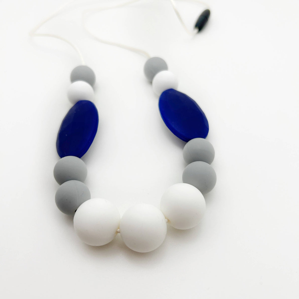 A silicone beaded necklace or teether featuring large white and medium gray round beads, accented by two large dark blue oval beads, on a white background.