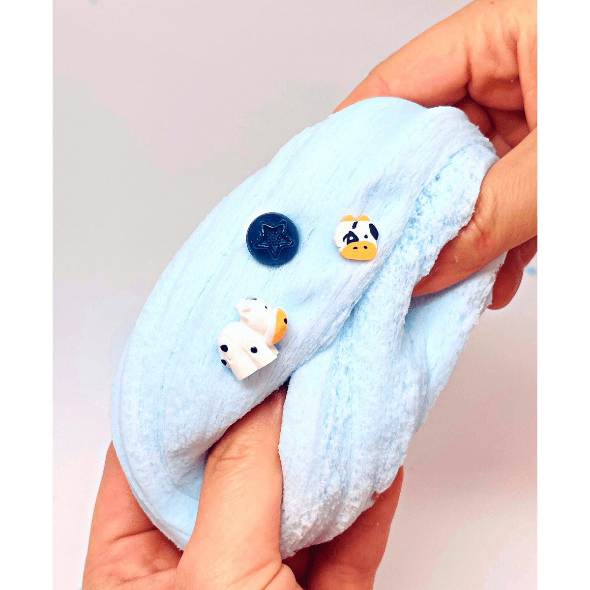 A close-up of a person's hands holding a light blue plush fabric item, possibly a small blanket, cushion, or pet toy, with three small decorative charms attached: a dark blue round charm, a white cow charm with black eyes, and a white duck charm with a yellow beak.
