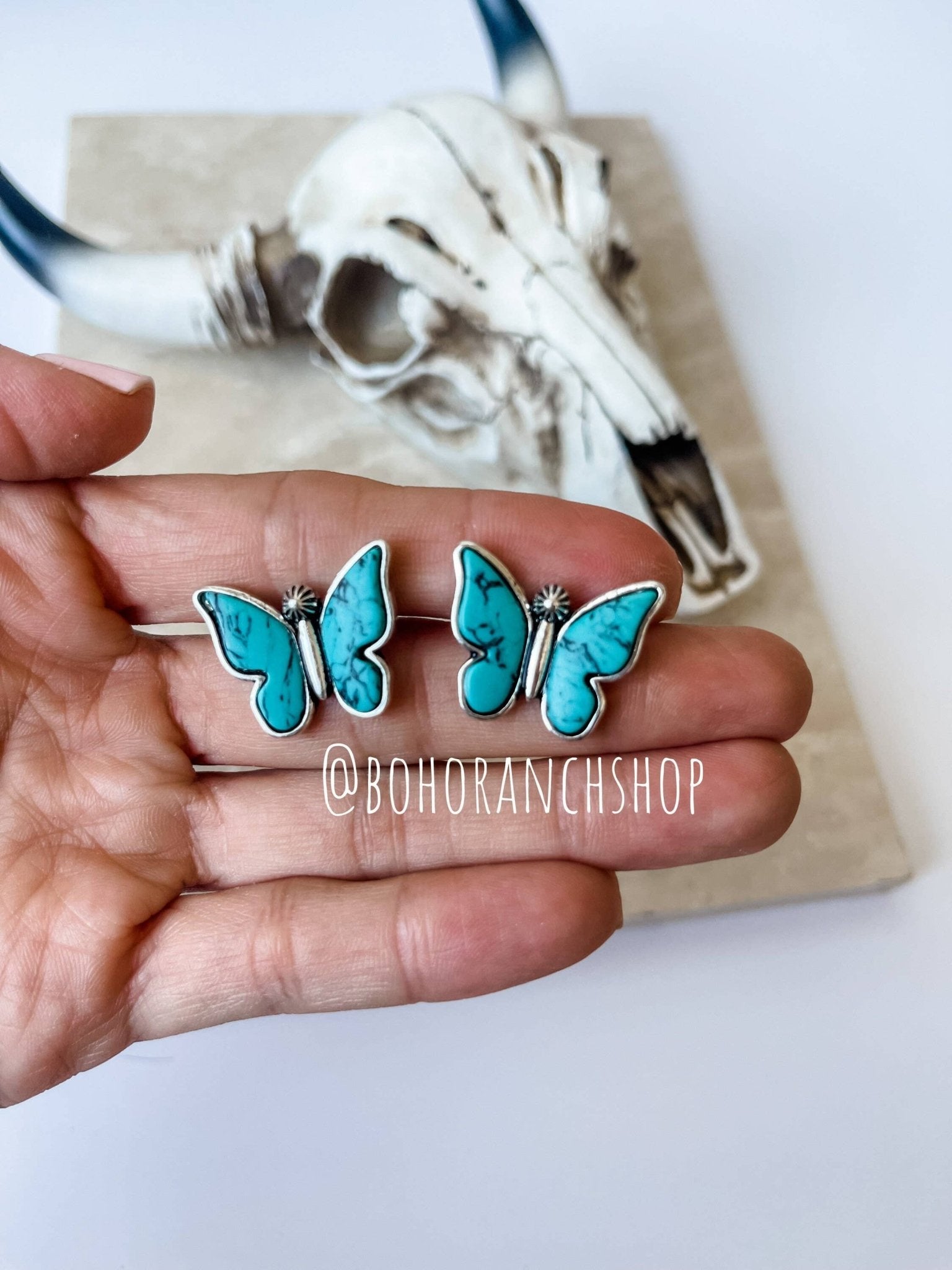 Turquoise butterfly earrings held in a hand with a skull and antlers in the background, branded @BohoRanchShop.