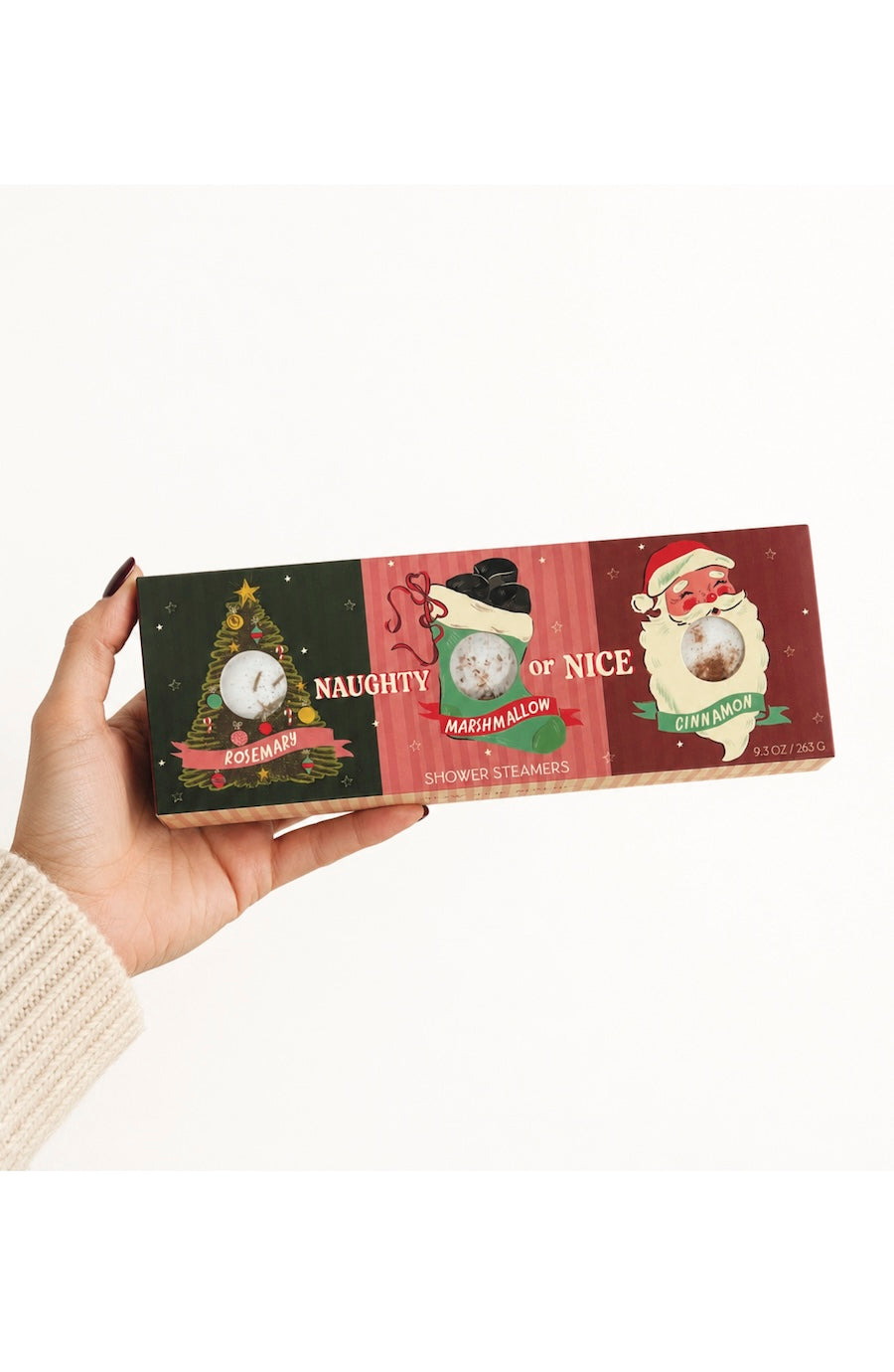 A hand holds a rectangular box of three holiday-themed mugs. The box features festive illustrations: a green panel with a Christmas tree and the word 'NAUGHTY', a red panel with a snowman and the phrase 'MADNESS ALLEY', and a dark red panel with Santa and the word 'GREATNESS'.