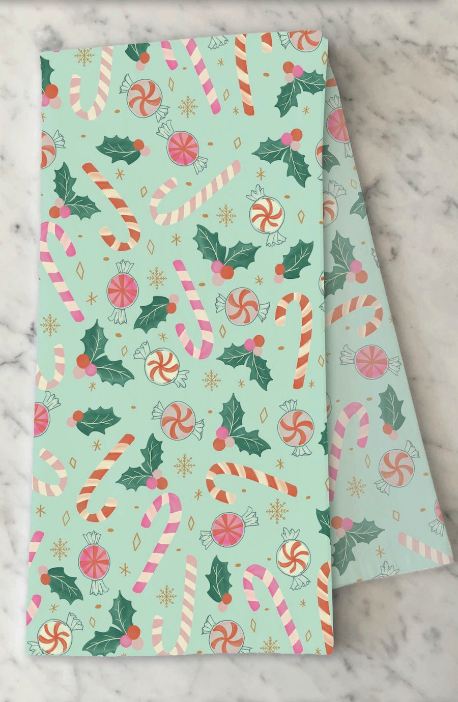 A folded sheet of wrapping paper with a festive Christmas pattern featuring candy canes, holly leaves, and peppermint candies on a mint green background, displayed on a white marble surface.