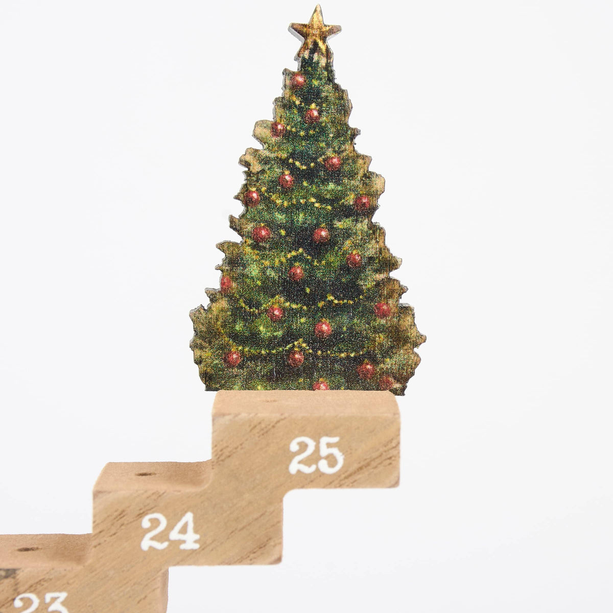 A Christmas tree advent calendar close-up showing a green Christmas tree with red ornaments and a gold star on top of wooden blocks numbered 25, 24, and 23 against a white background.