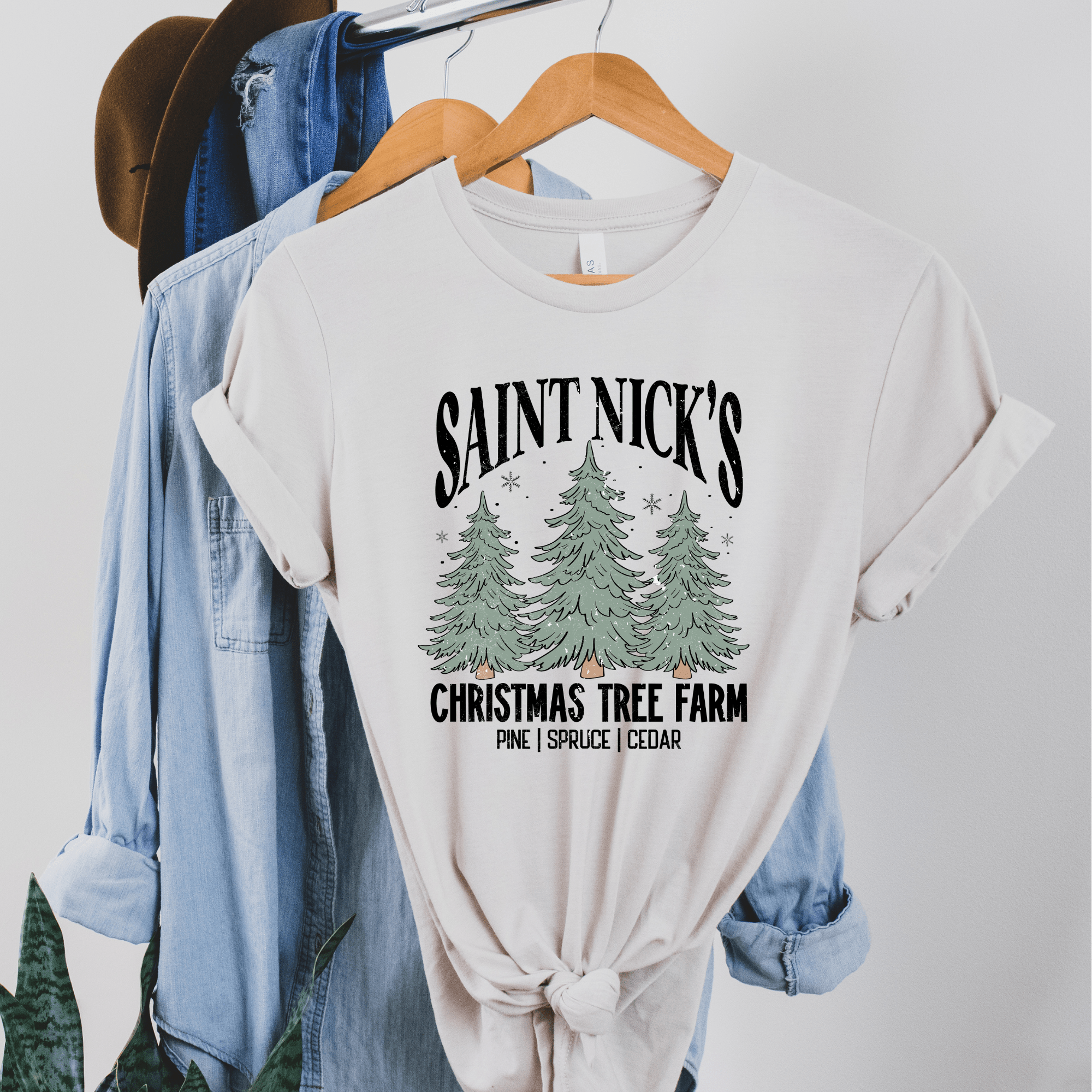 White t-shirt with 'Saint Nick's Christmas Tree Farm' design on a hanger with jeans and a hat.