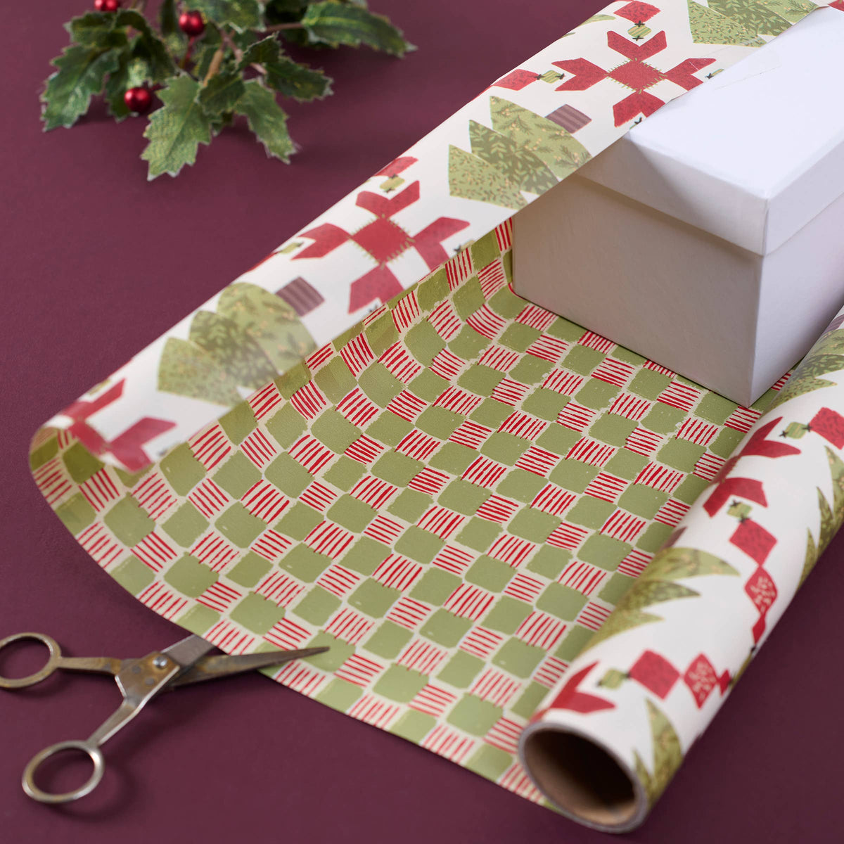 Holiday-themed wrapping paper with a green and red geometric pattern, shown partially unrolled over a white gift box with metal scissors and holly in the background.