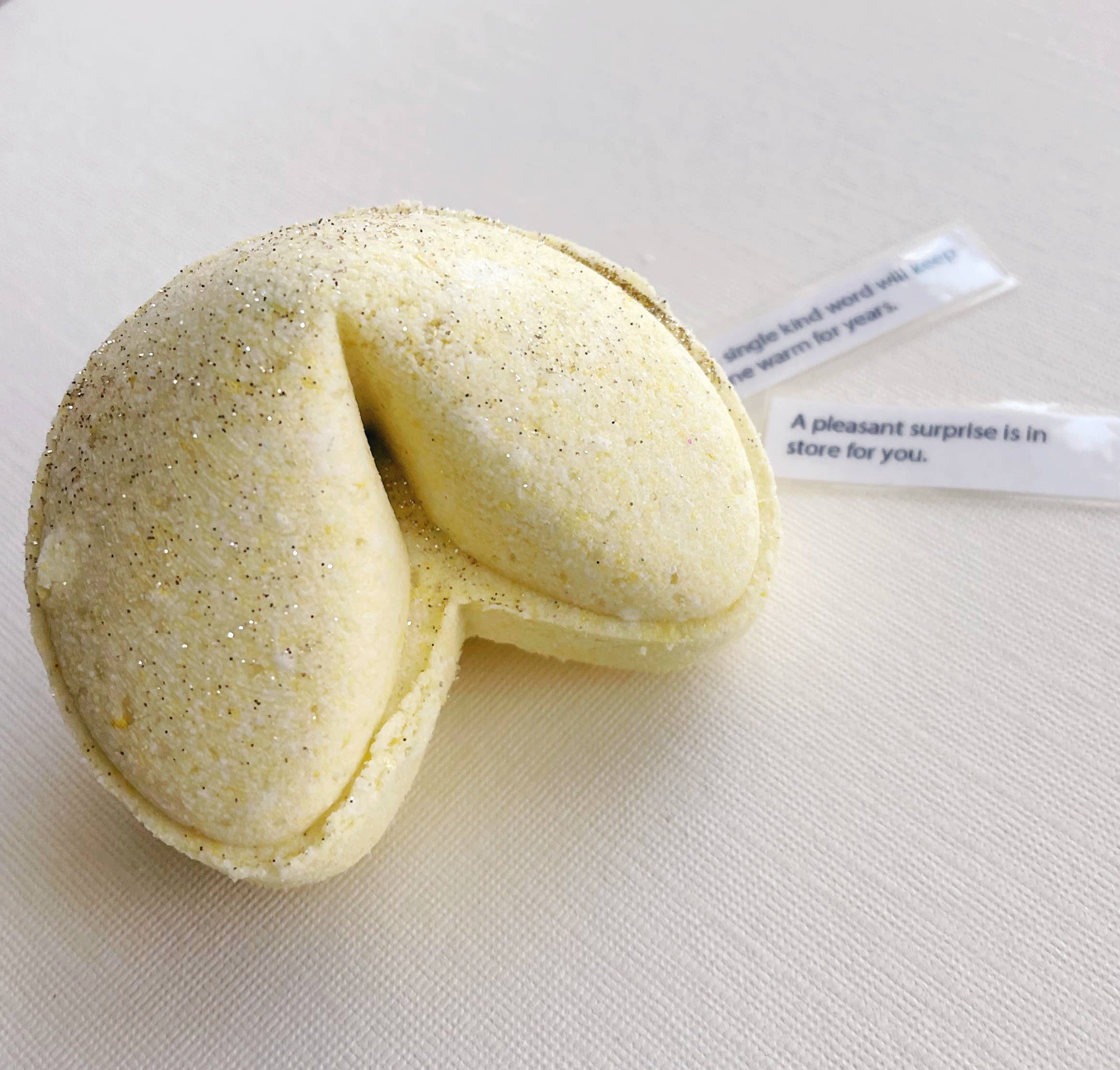 A bath bomb shaped like a fortune cookie, with a label that reads 'A pleasant surprise is in store for you.'