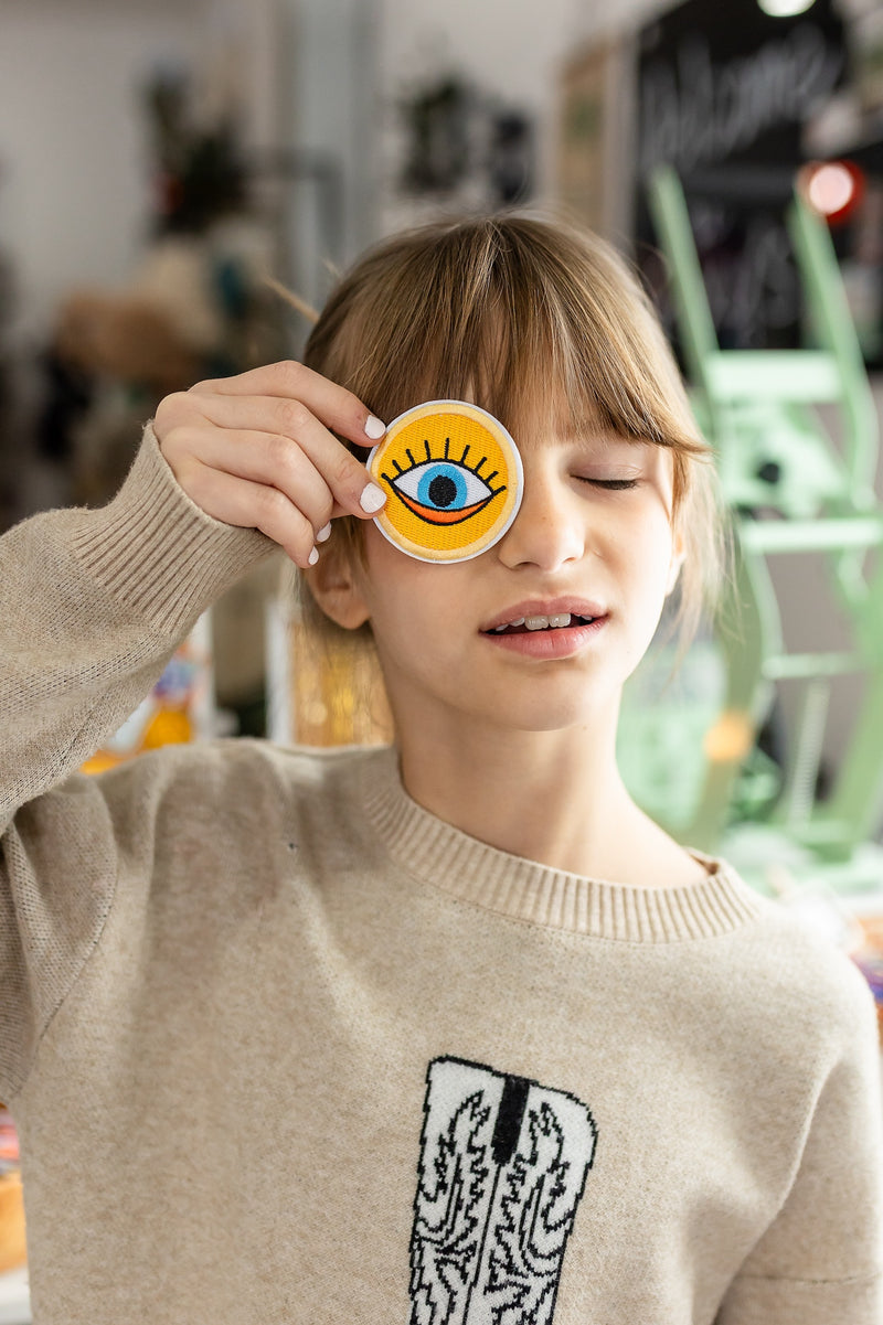 Person wearing a beige sweater with a graphic design, holding up a sticker with an eye symbol.