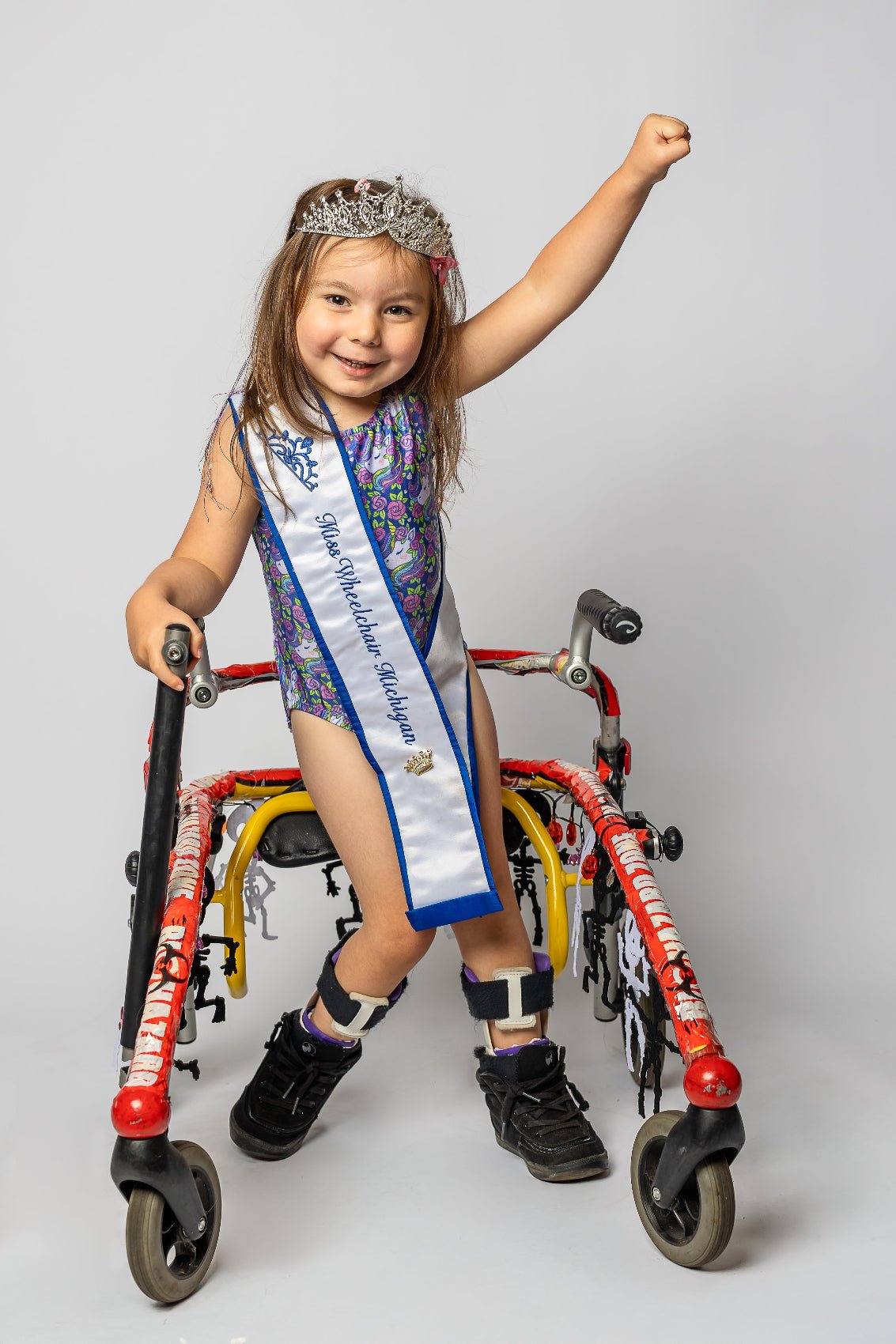 Young girl with a walker wearing a sash and tiara on a plain background