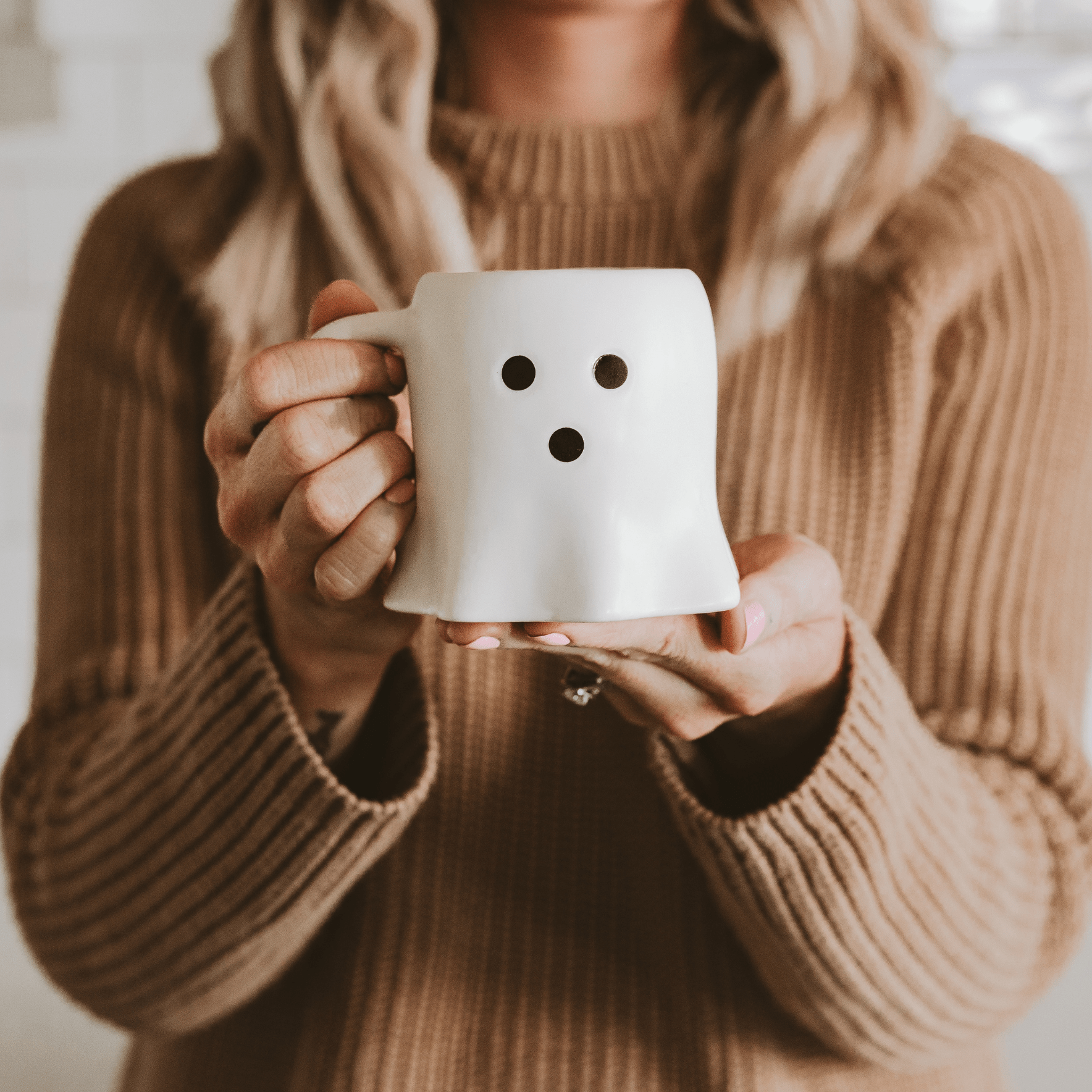 Person holding a white mug with a ghost face design, wearing a brown sweater.