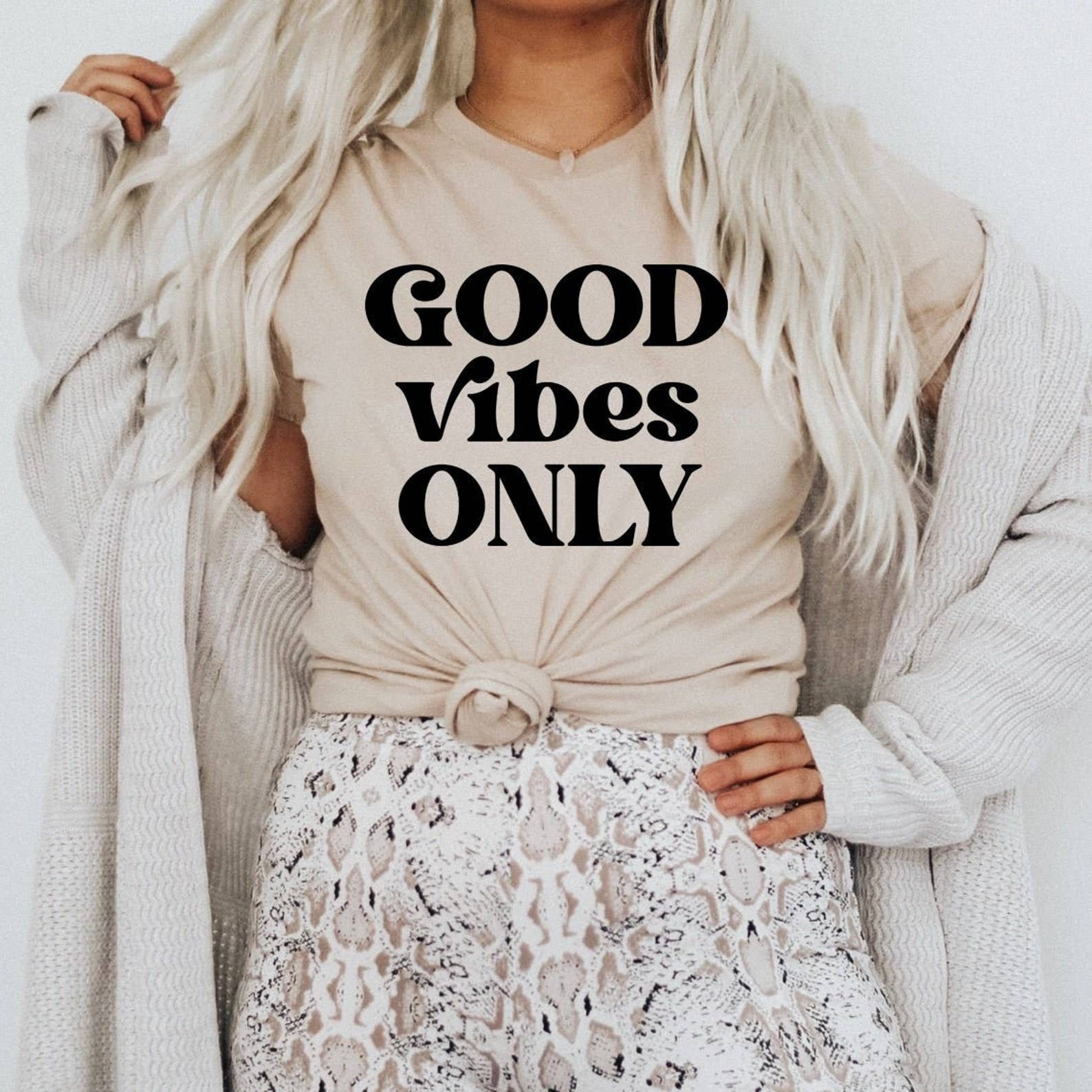 Person wearing a beige shirt with 'GOOD VIBES ONLY' text, white lace pants, and a light gray cardigan.