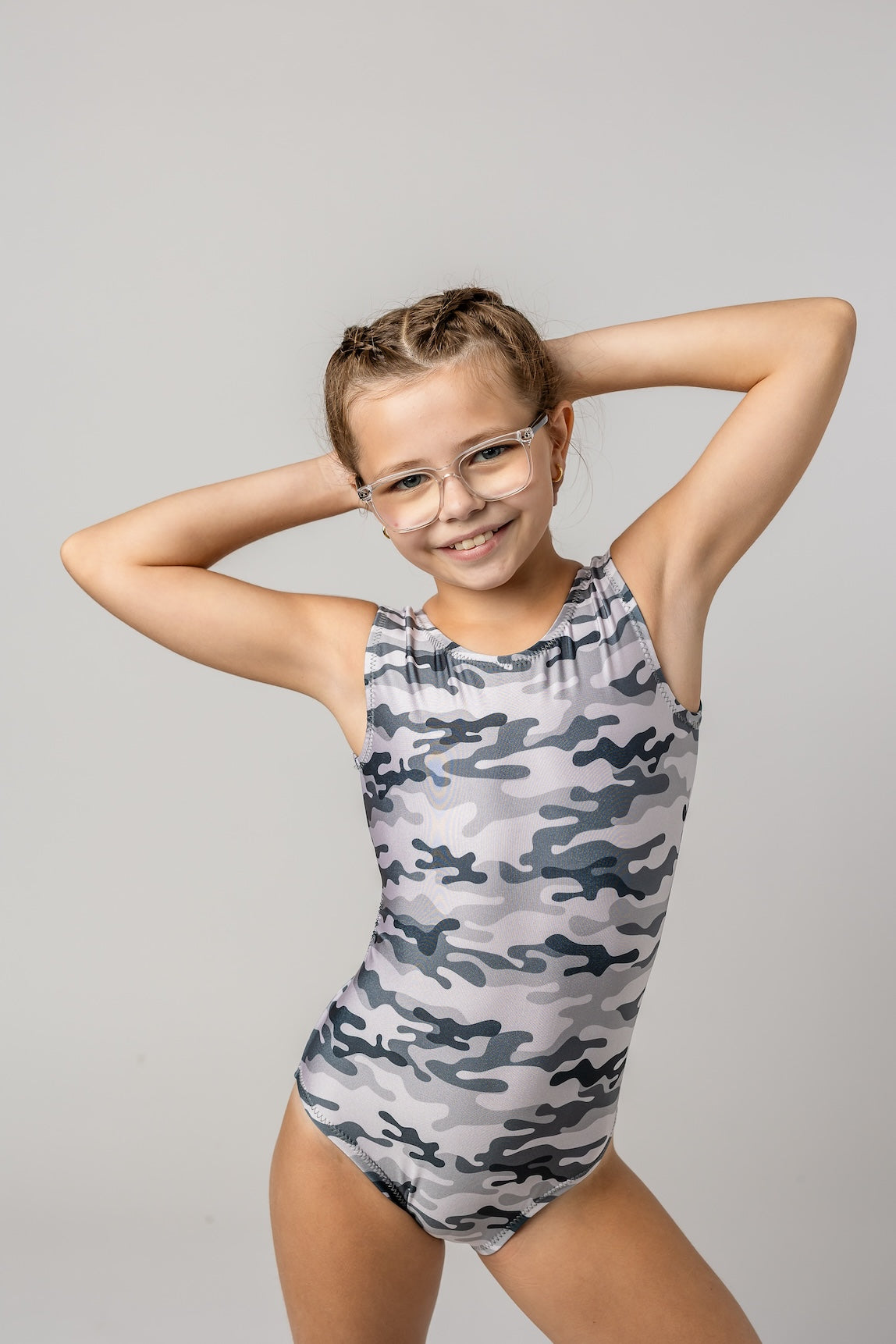 A young girl with glasses posing in a gray and white camouflage print leotard against a light gray background.