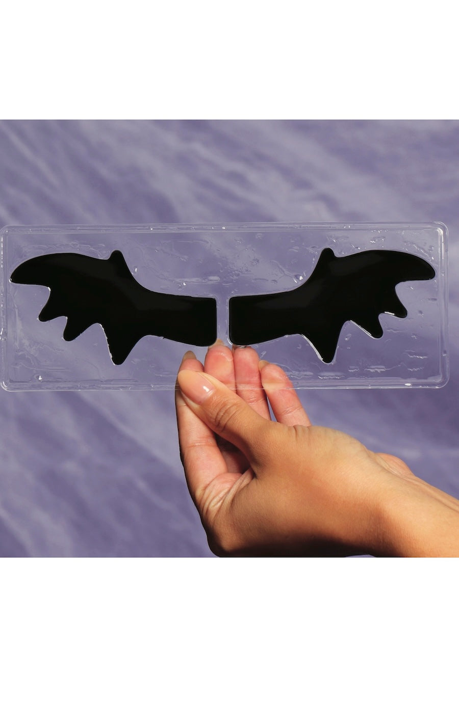 A hand holding a clear plastic mold or tray with two black bat-shaped pieces, shown against a purple textured background.