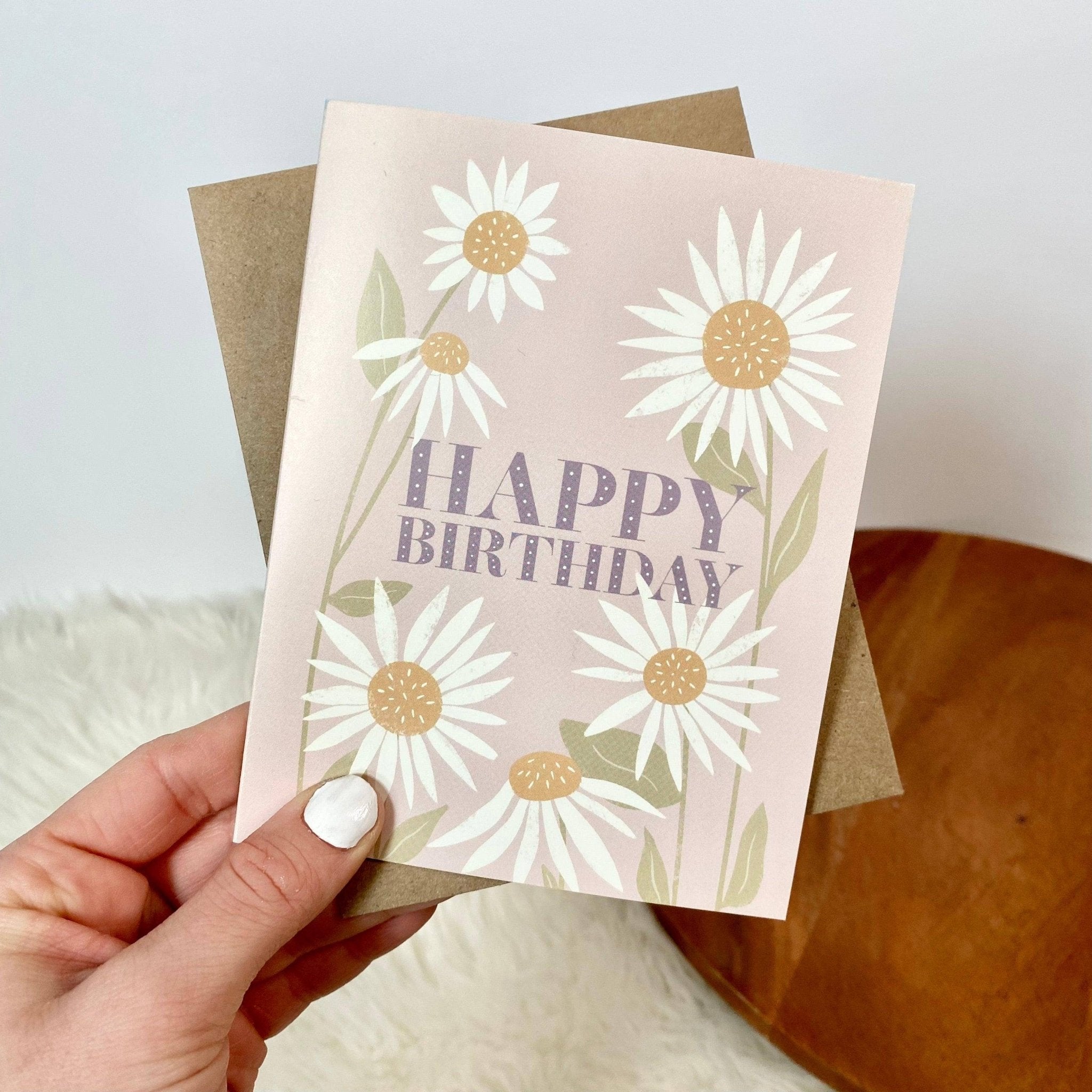 Pink 'Happy Birthday' card with daisy floral design and brown envelope held in hand.