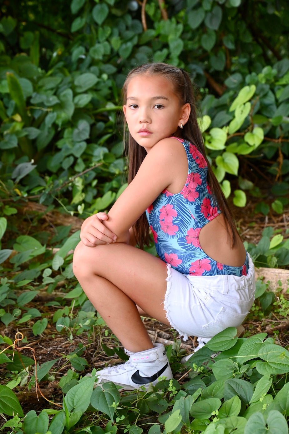 Young gymnast sitting on the ground with green foliage in a turquoise blue leotard with large pink hibiscus flower print