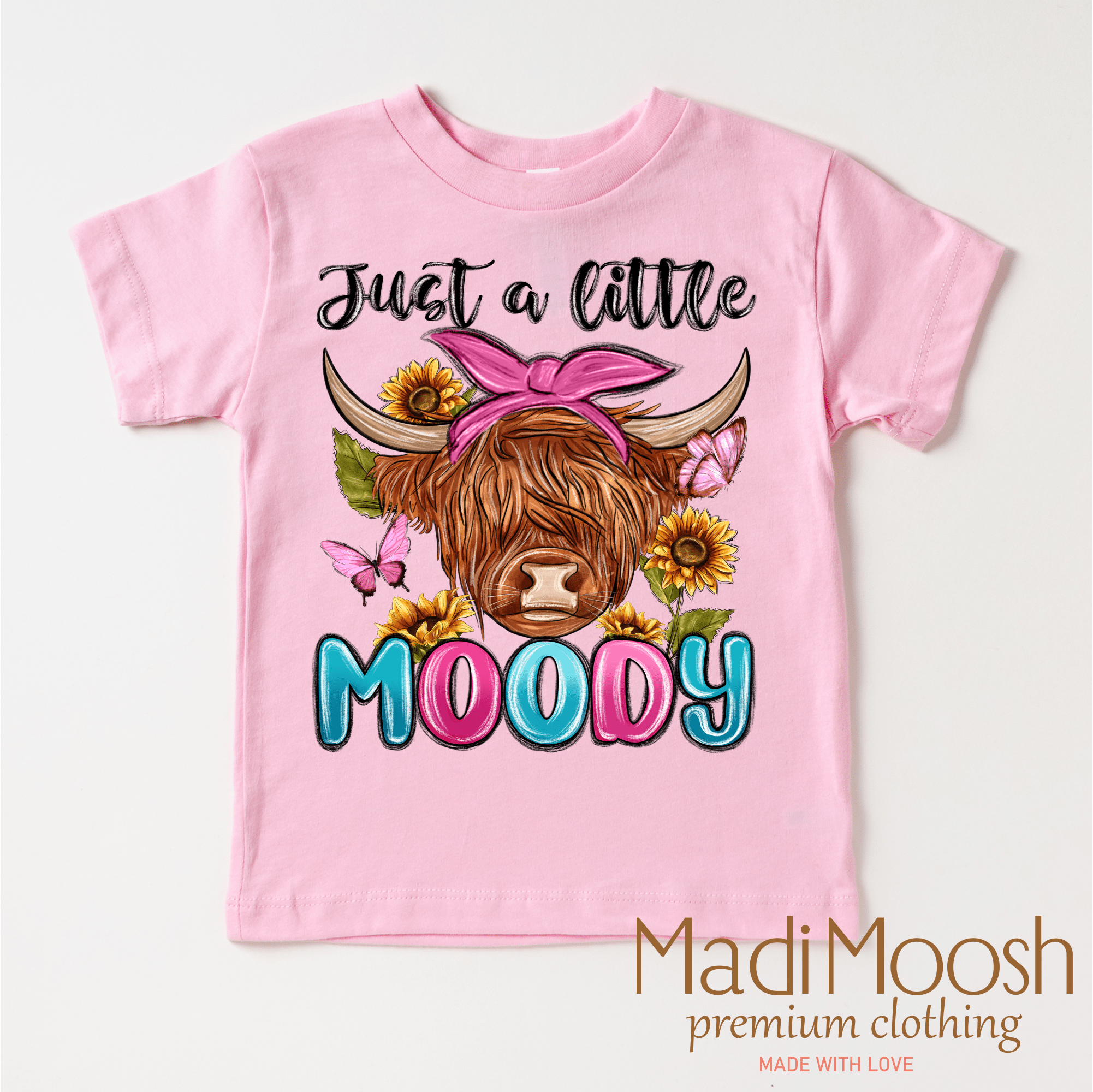 Pink t-shirt with a cow graphic and 'Just a little Moody' text on a white background, featuring the brand 'Madi Moosh'.