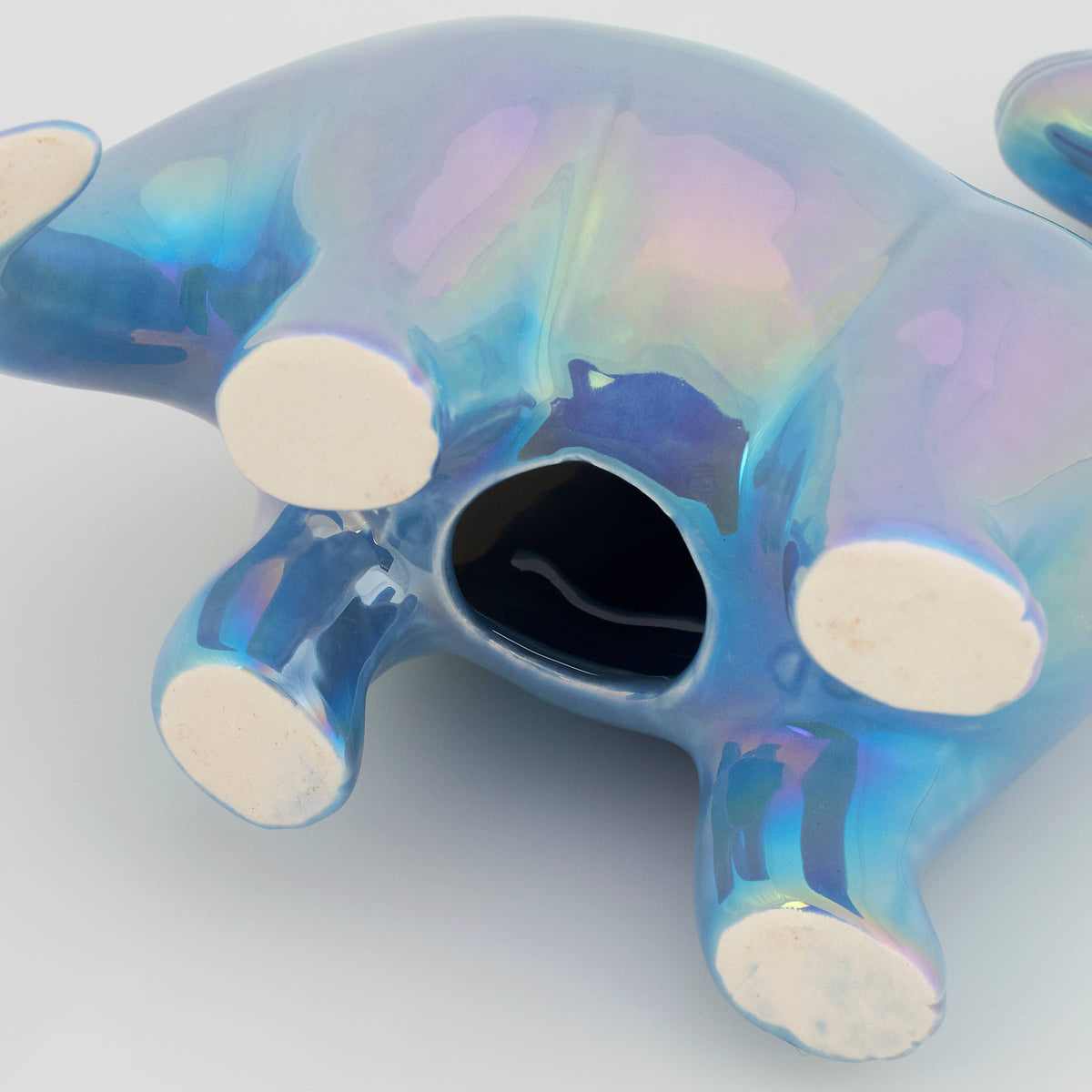 A glossy iridescent blue ceramic animal figurine, shown from a bottom angle with its feet and back visible. The surface has a holographic, rainbow-like sheen shifting between blue, purple, and pink, with matte off-white circular feet and a dark opening in the center.