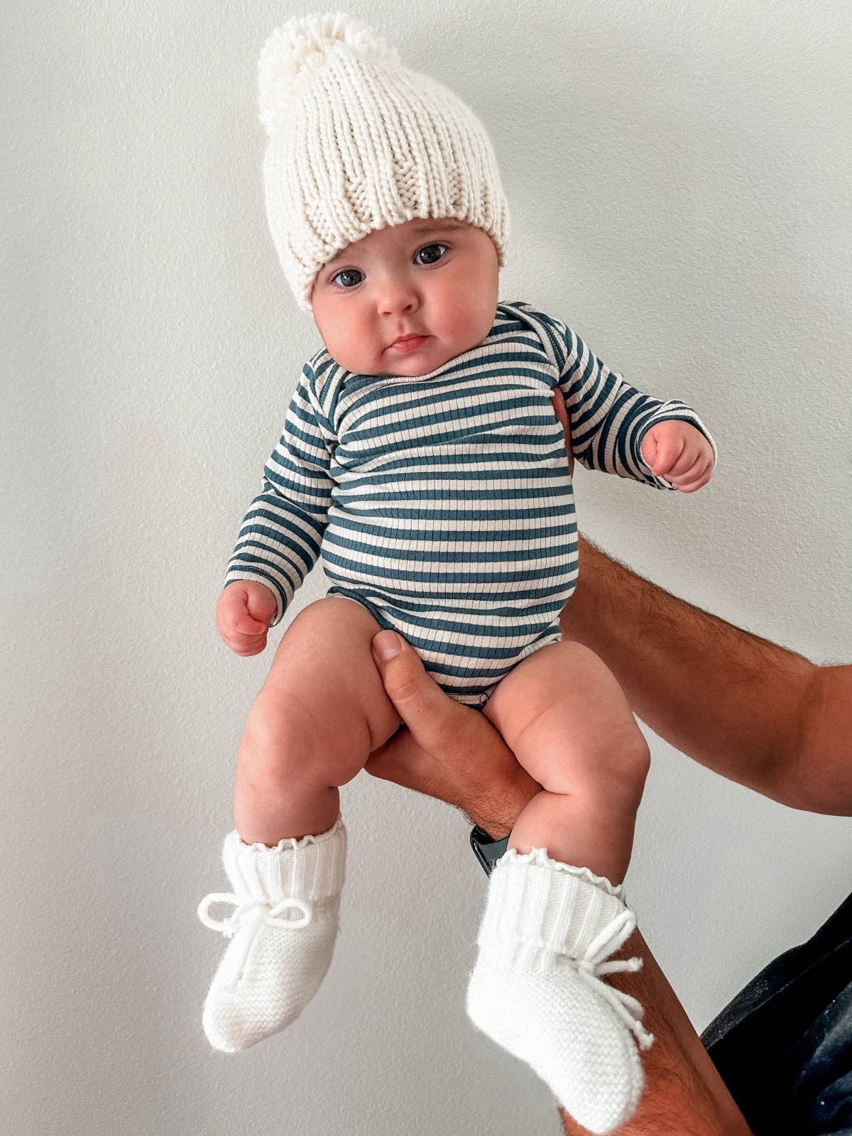Baby in striped onesie and white knit hat and booties being held against a plain background