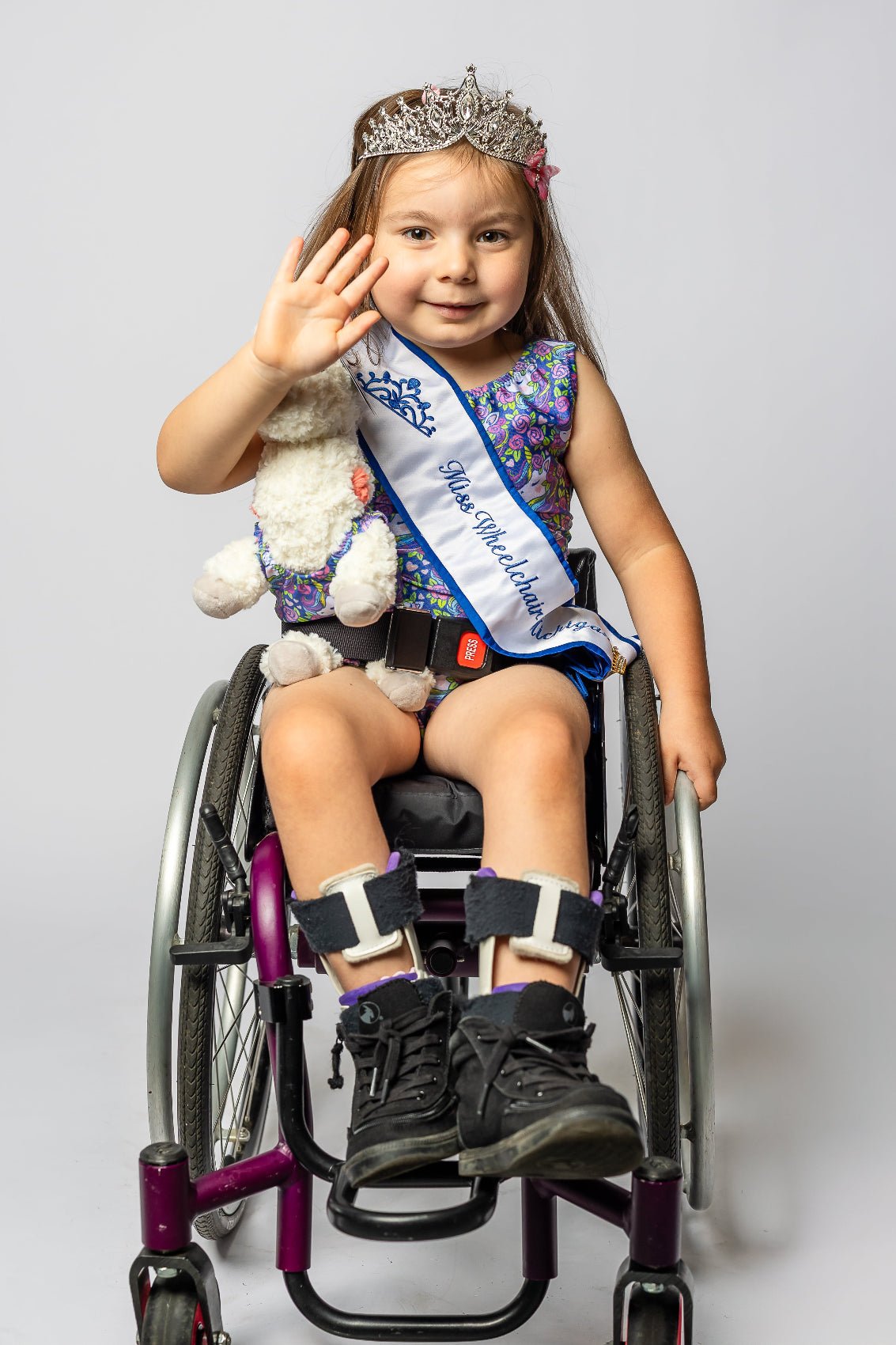 Young girl in a wheelchair wearing a sash and tiara, waving at the camera, in her Foxy's Tumble Tot Set with a unicorn pattern