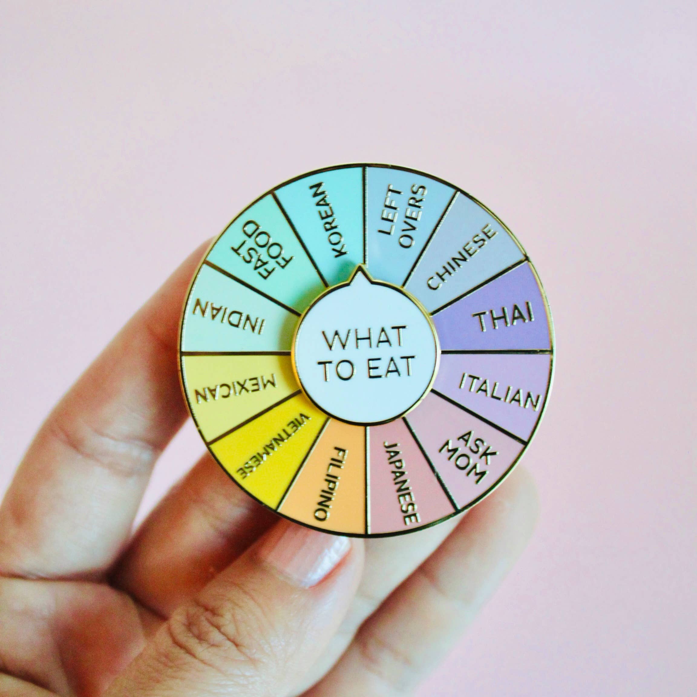 A hand holding a round, hard enamel pin with different food genres like Indian, Korean, Chinese, Thai, Italian, and Ask Mom, each labeled with their respective names.