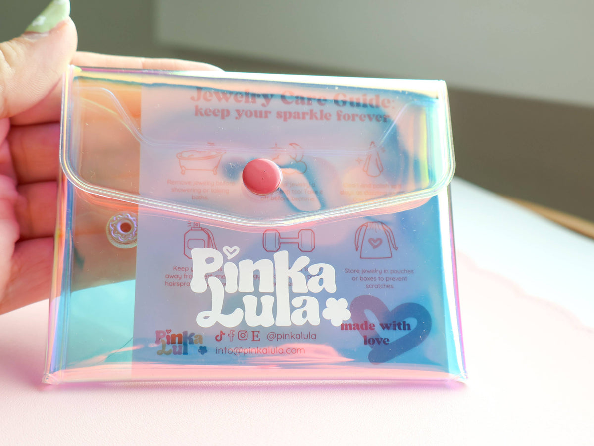 A hand holding a holographic iridescent pouch with a pink snap button and a printed jewelry care guide insert. The pouch features the brand name 'Pinka Lula' in white script with a small heart.